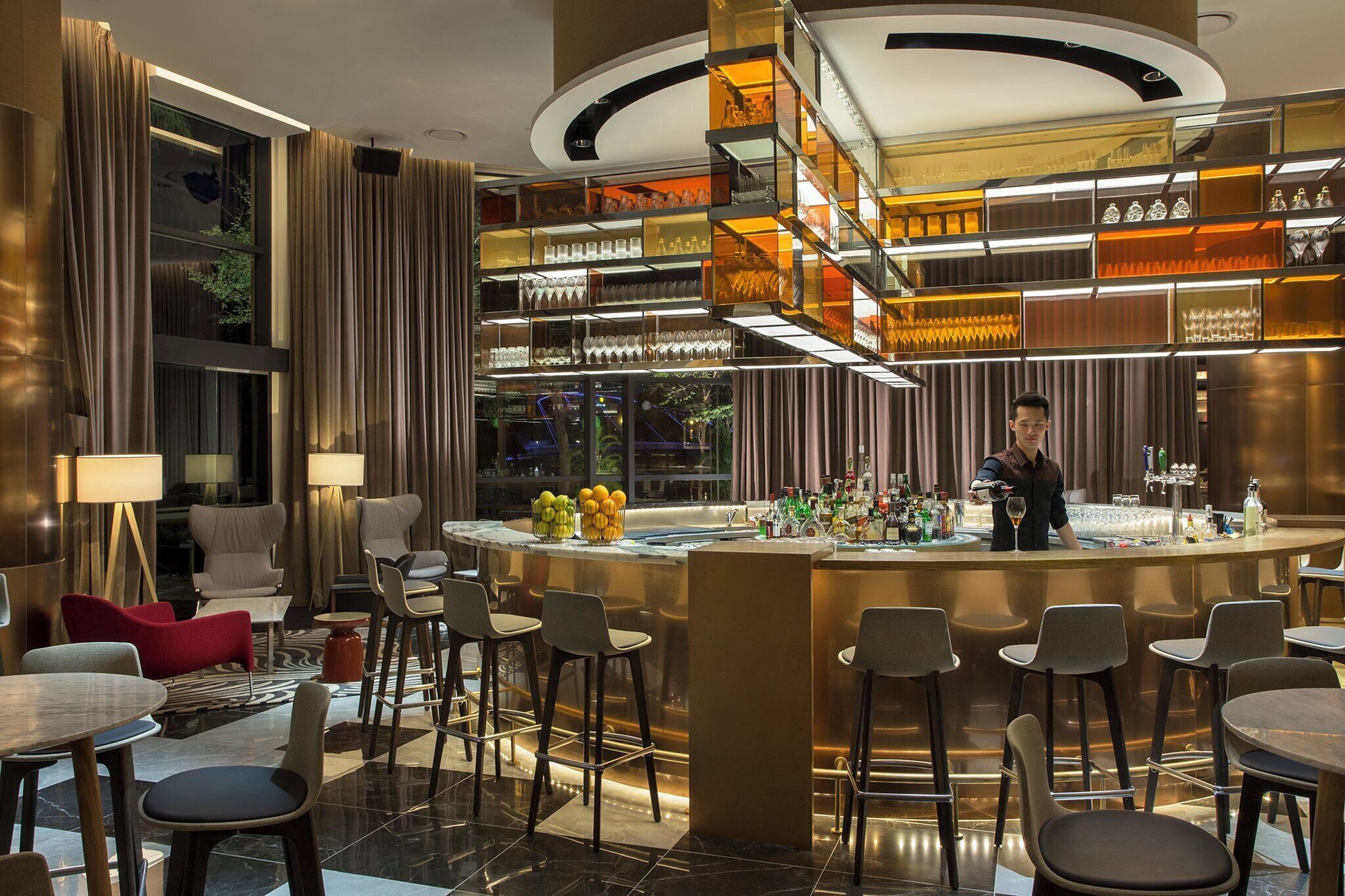 A modern bar area with a bartender, bar stools, and a variety of glassware displayed on shelves.