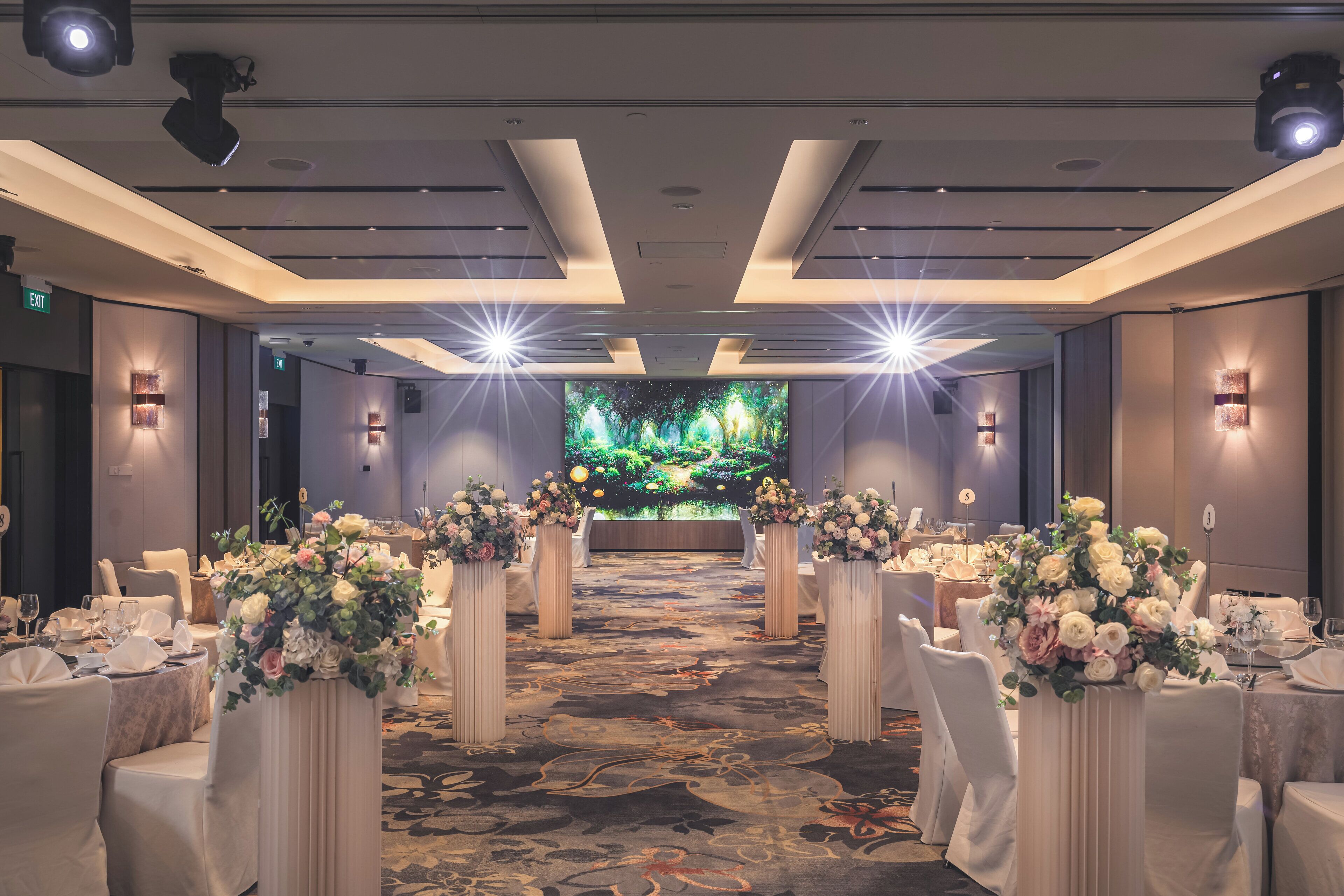 A banquet hall with a long table set for a formal event, flanked by tall flower arrangements.