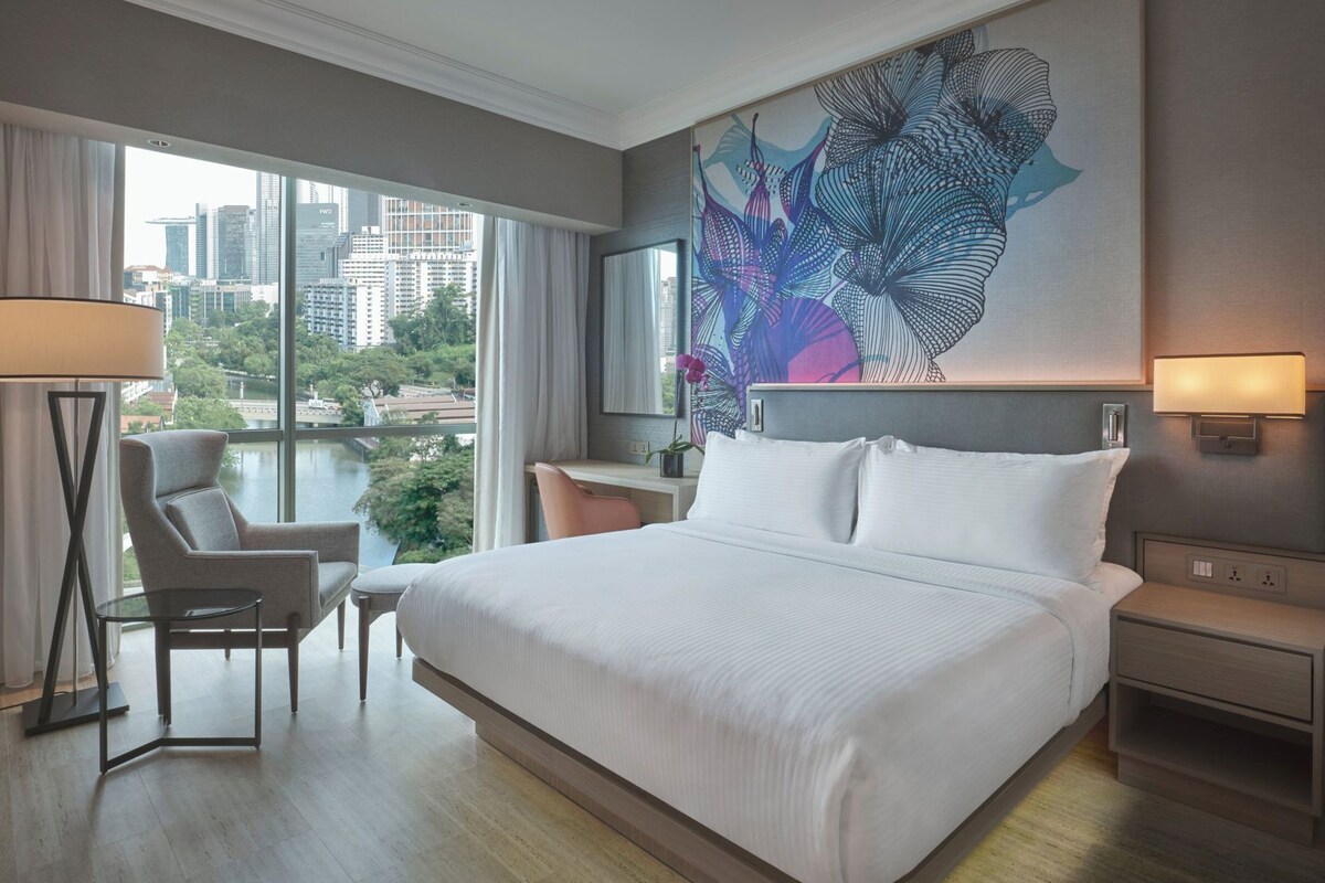 A modern hotel room with a large bed, a desk, a chair, and a view of a cityscape through large windows.