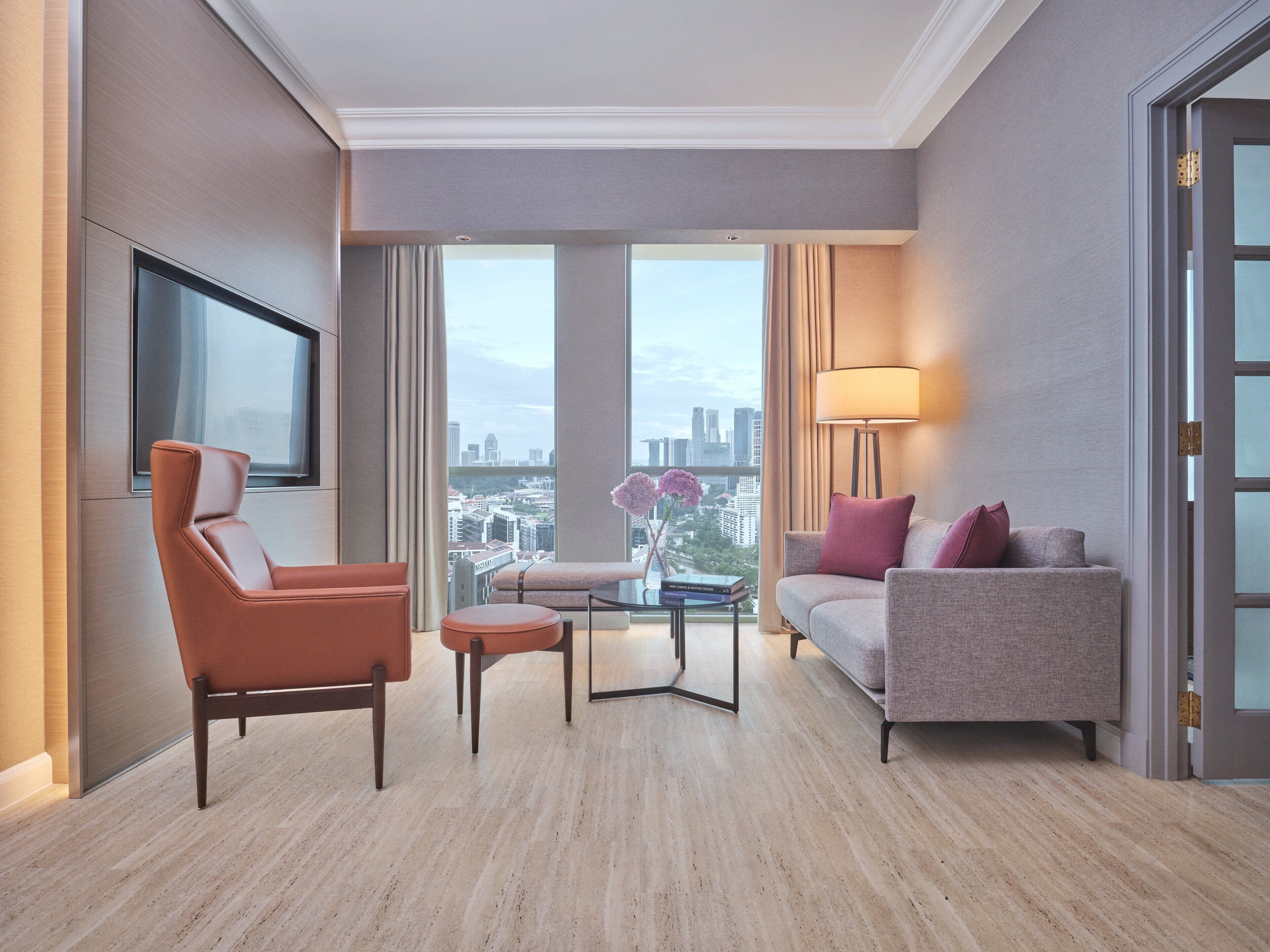 Ein modernes Hotelzimmer mit großem Fenster und Blick auf die Stadt, einem Sofa, einem Sessel und einem kleinen Tisch mit Lampe.
