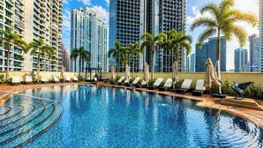 Una piscina con sillones de playa y sombrillas, rodeada de edificios altos y palmeras.