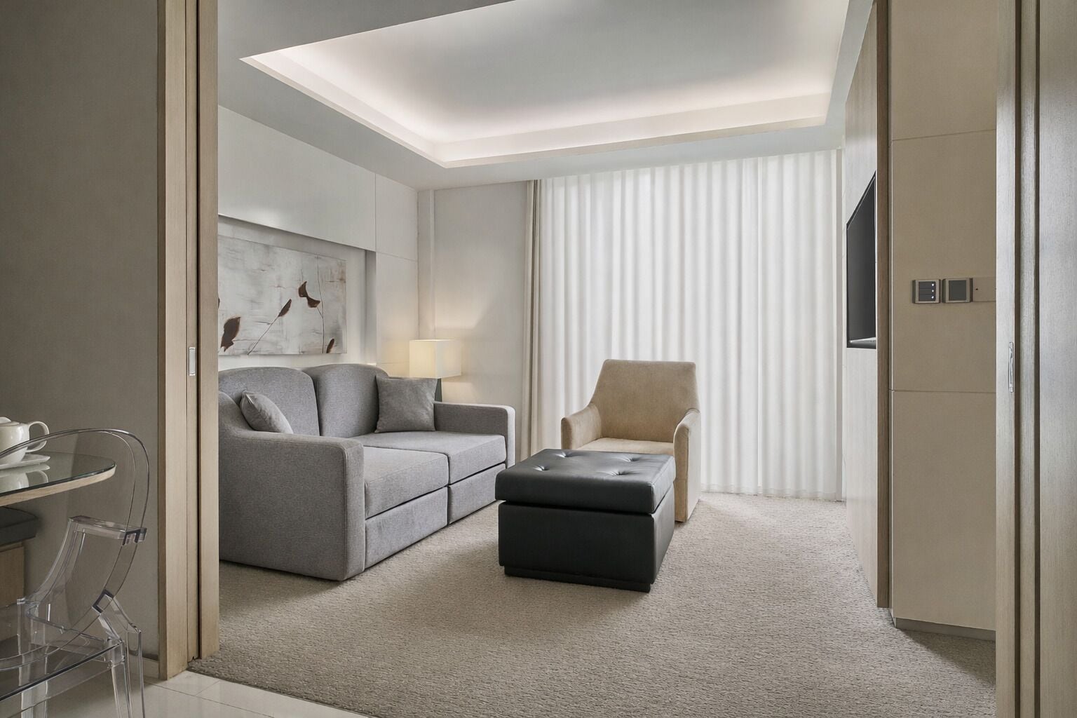 A modern hotel room with a grey sofa, a black ottoman, a beige armchair, a glass coffee table, and a wall-mounted TV.