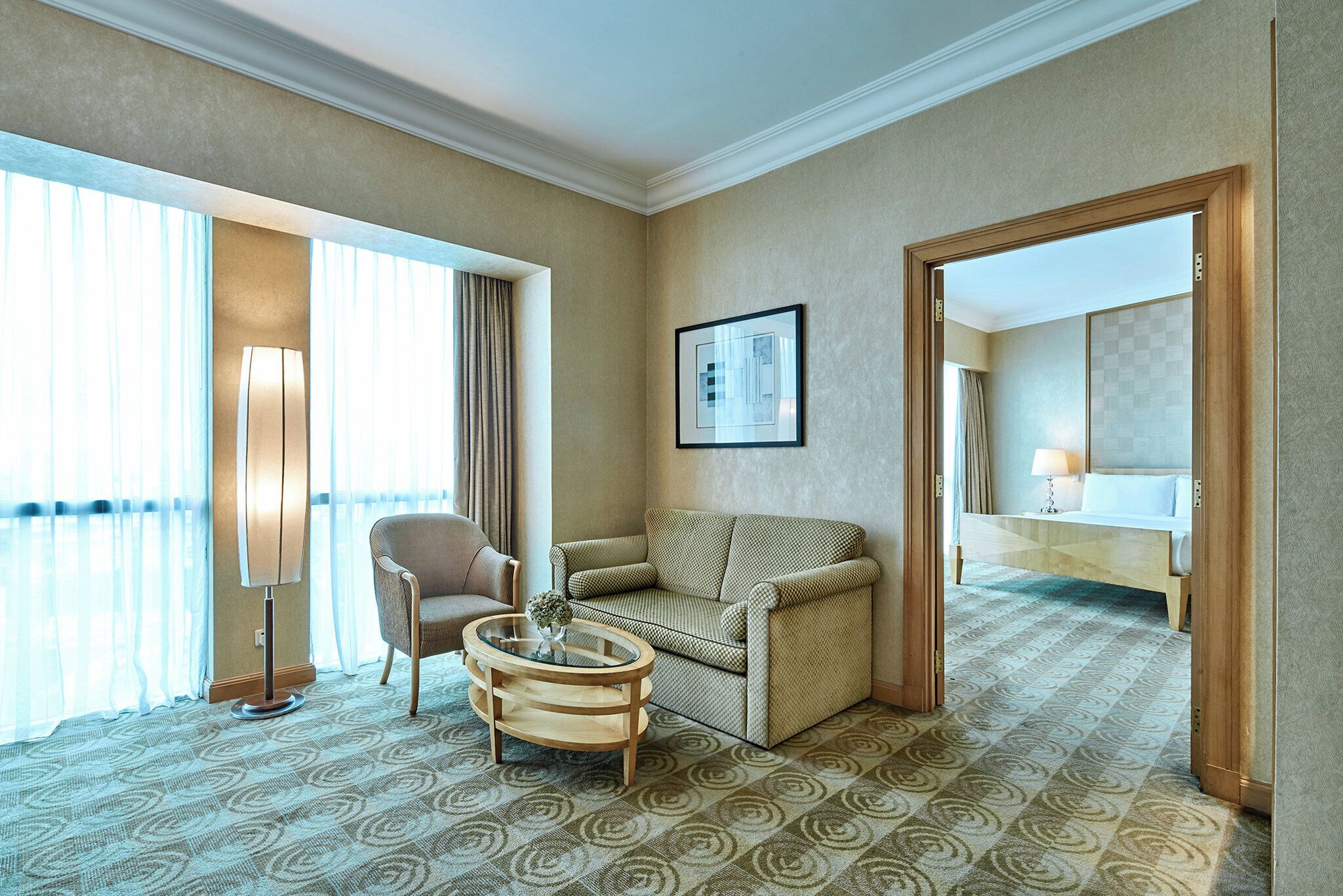 A hotel room with a sofa, armchair, and a small round table. There is a large window with curtains, a framed picture on the wall, and a mirror in the corner.