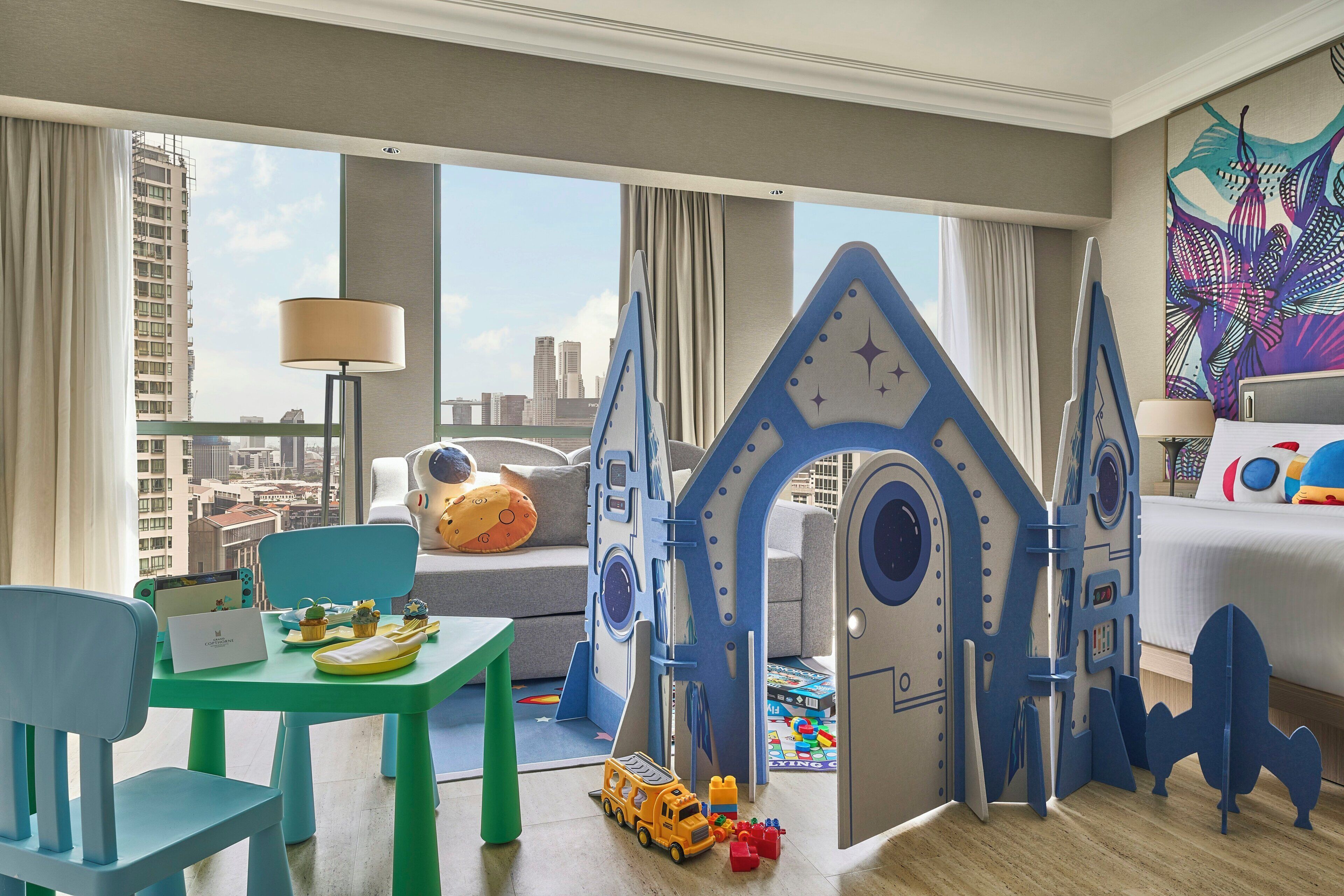 A children's play area with a playhouse, a table with toys, and a view of the city through large windows.