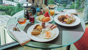 Una mesa de desayuno con diversos platos y bebidas, una cafetera y una vista panorámica de la ciudad a través de amplios ventanales.