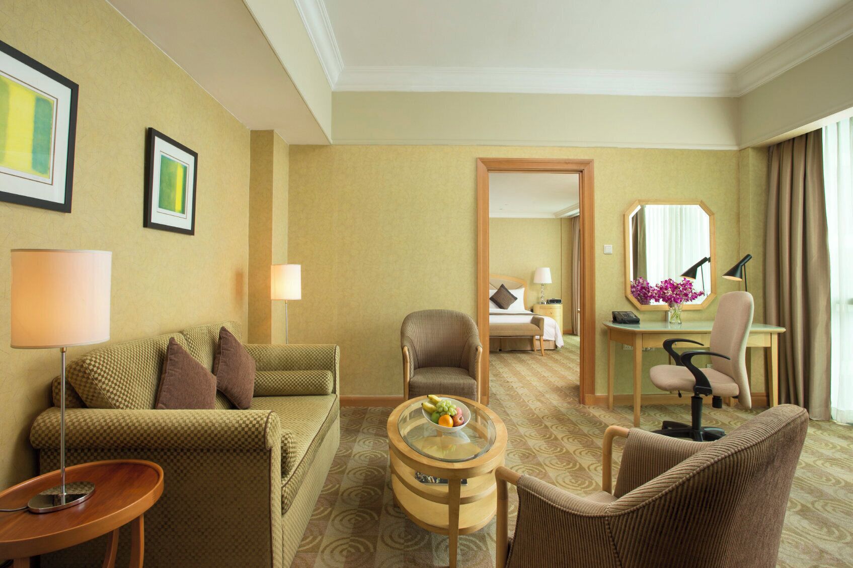 A hotel room with a sofa, armchair, round coffee table, and a desk with a chair. There is a bed with a nightstand and lamp, and a mirror on the wall.