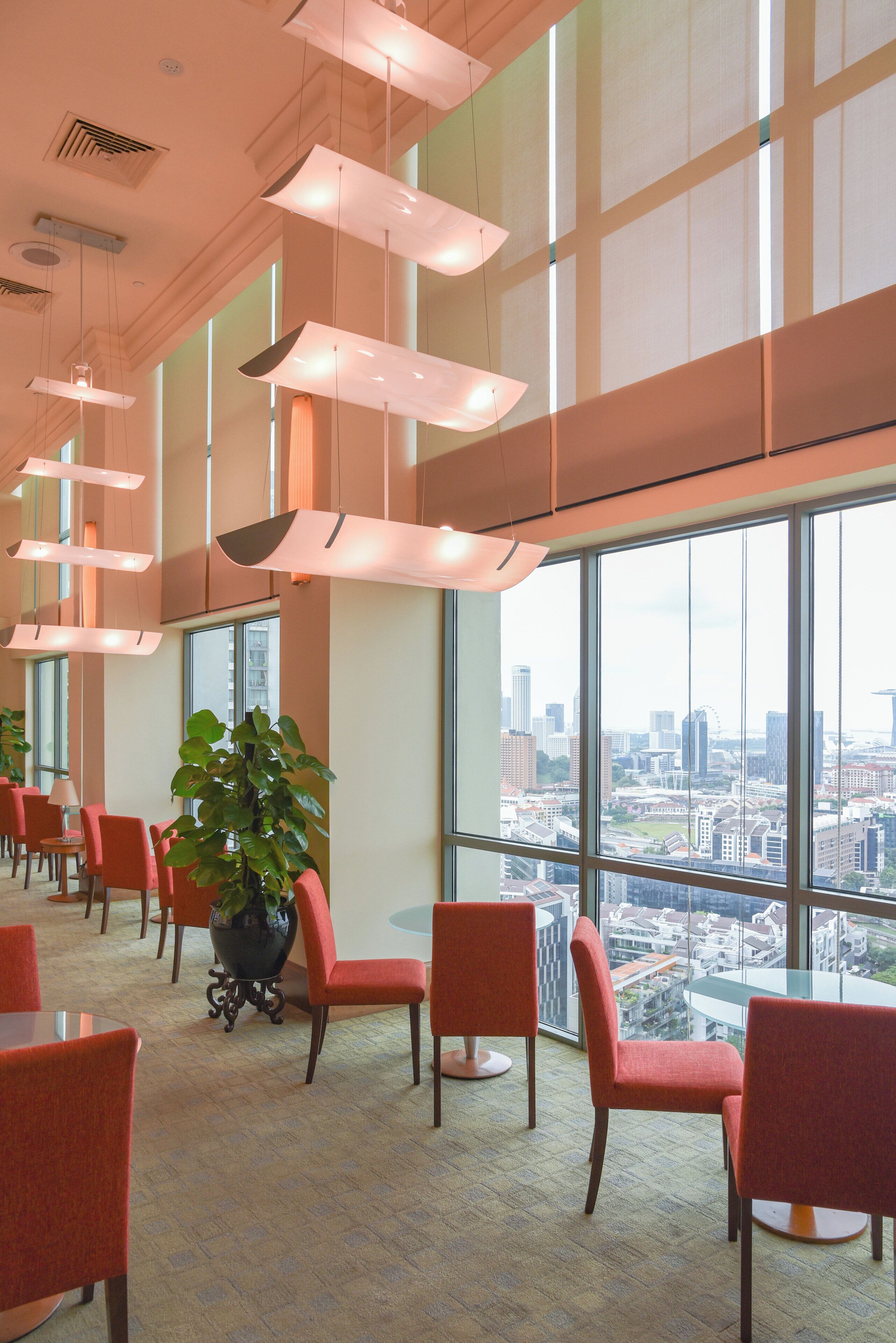 A spacious lobby with large windows offering a city view, modern pendant lighting, and red upholstered chairs.