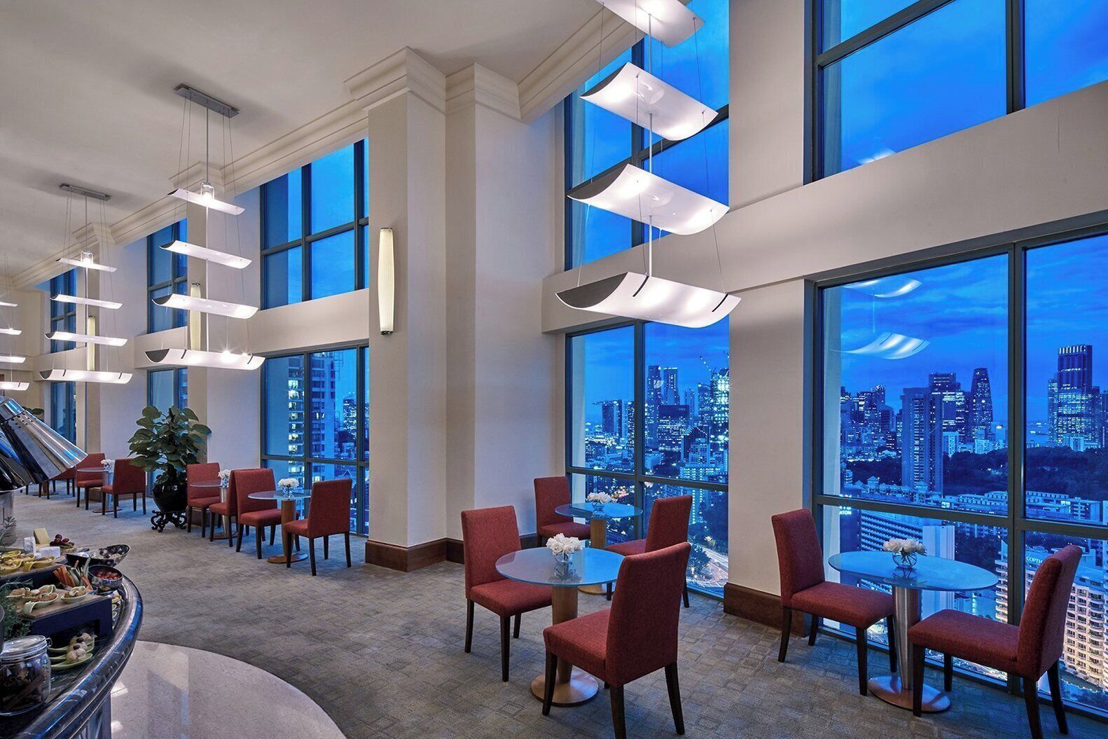 A modern hotel lobby with large windows offering a city view, red upholstered chairs, and a buffet area.