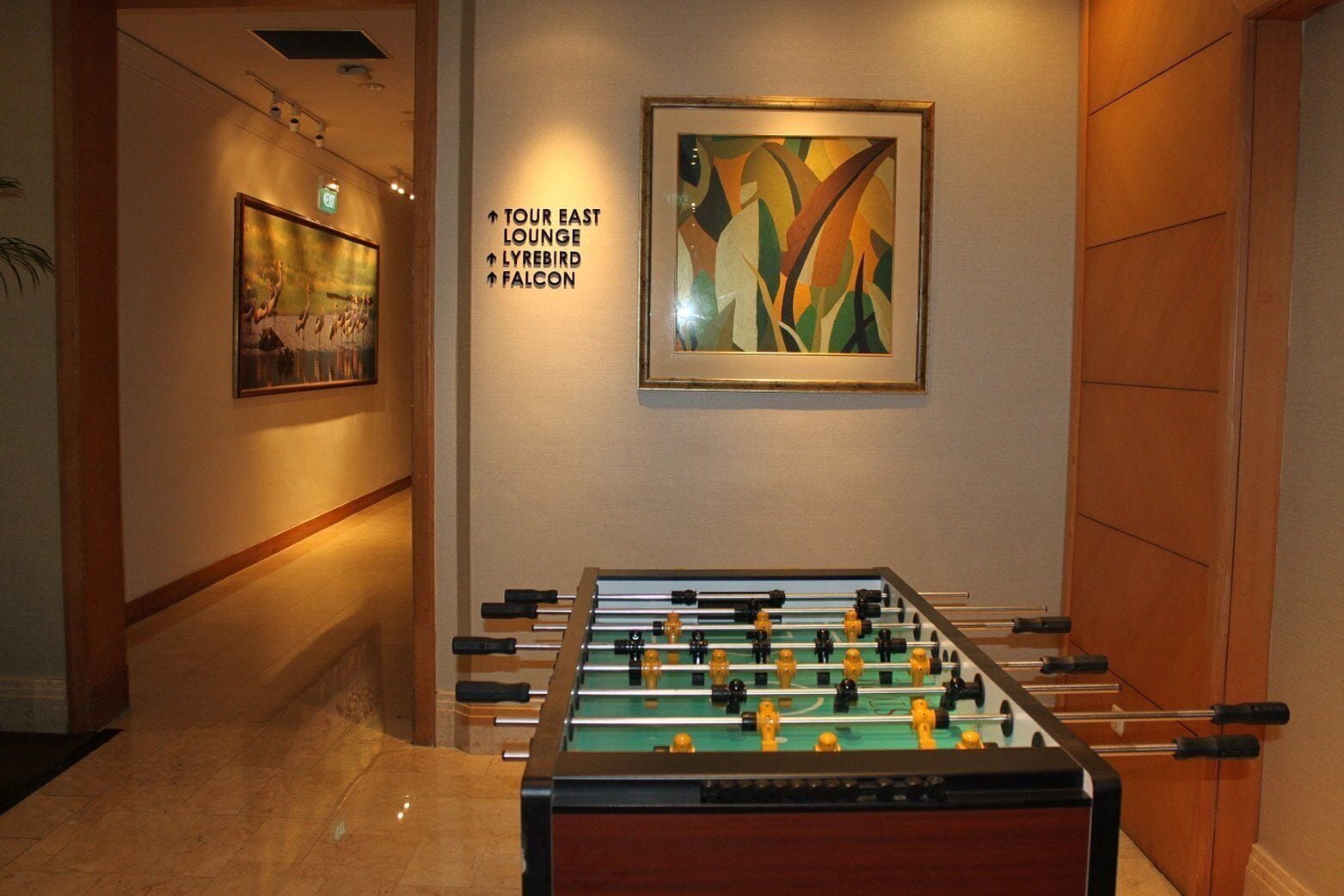 A hotel lobby with a foosball table, directional signs to the east lounge, lyrebird, and falcon, and a framed abstract painting.