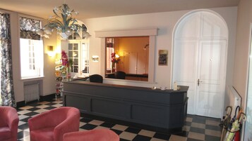 Un área de recepción con piso a cuadros, un mostrador de recepción, un espejo grande, una puerta blanca y una planta.