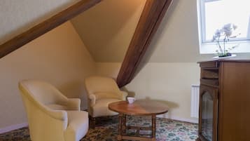 Een kamer met een schuine kap, twee crèmekleurige stoelen, een klein houten tafeltje, een kast met spiegel en een raam met een orchideeënplant.
