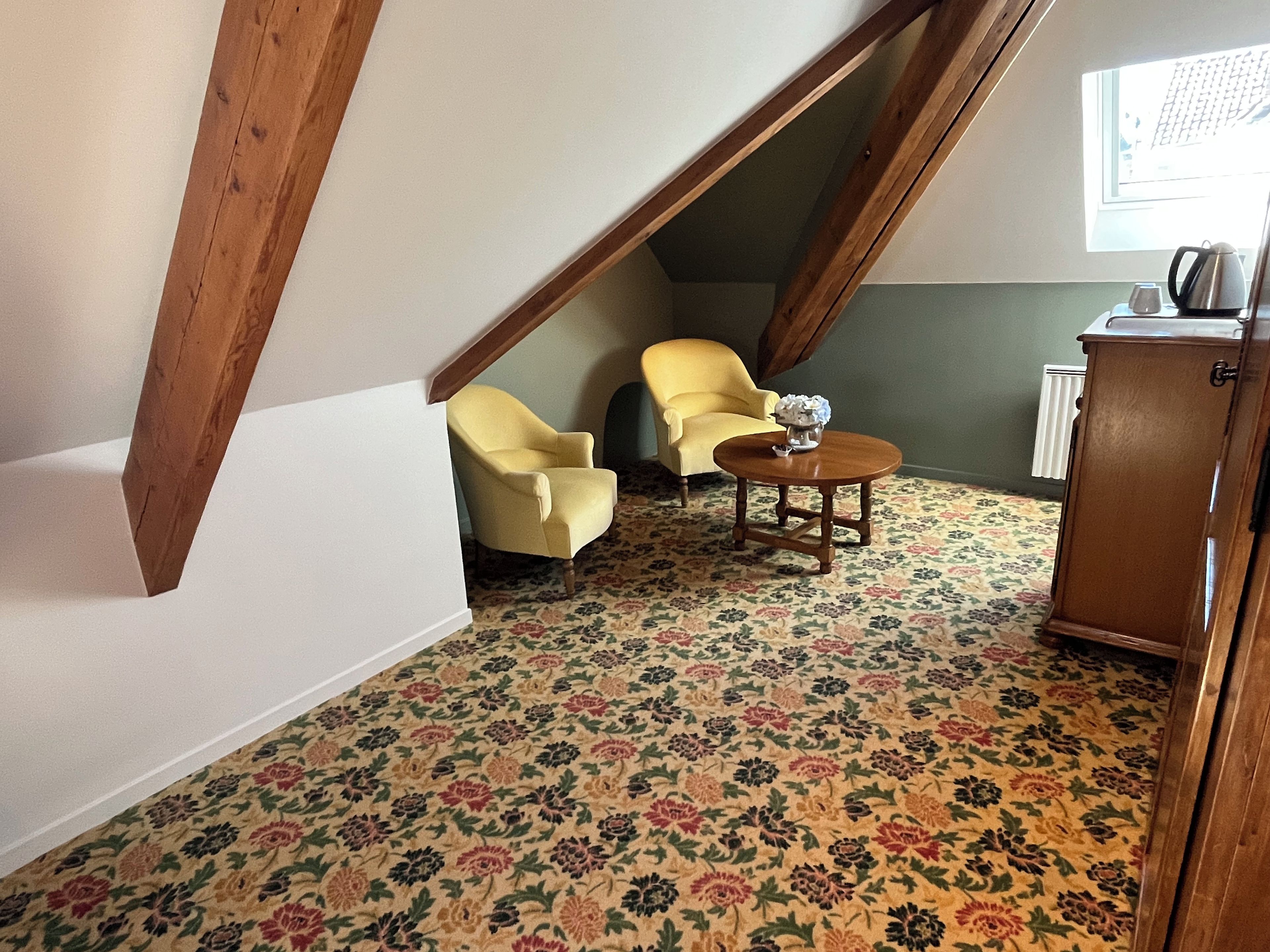 A room with a sloped ceiling, two yellow armchairs, a small wooden table with a vase, and a radiator.