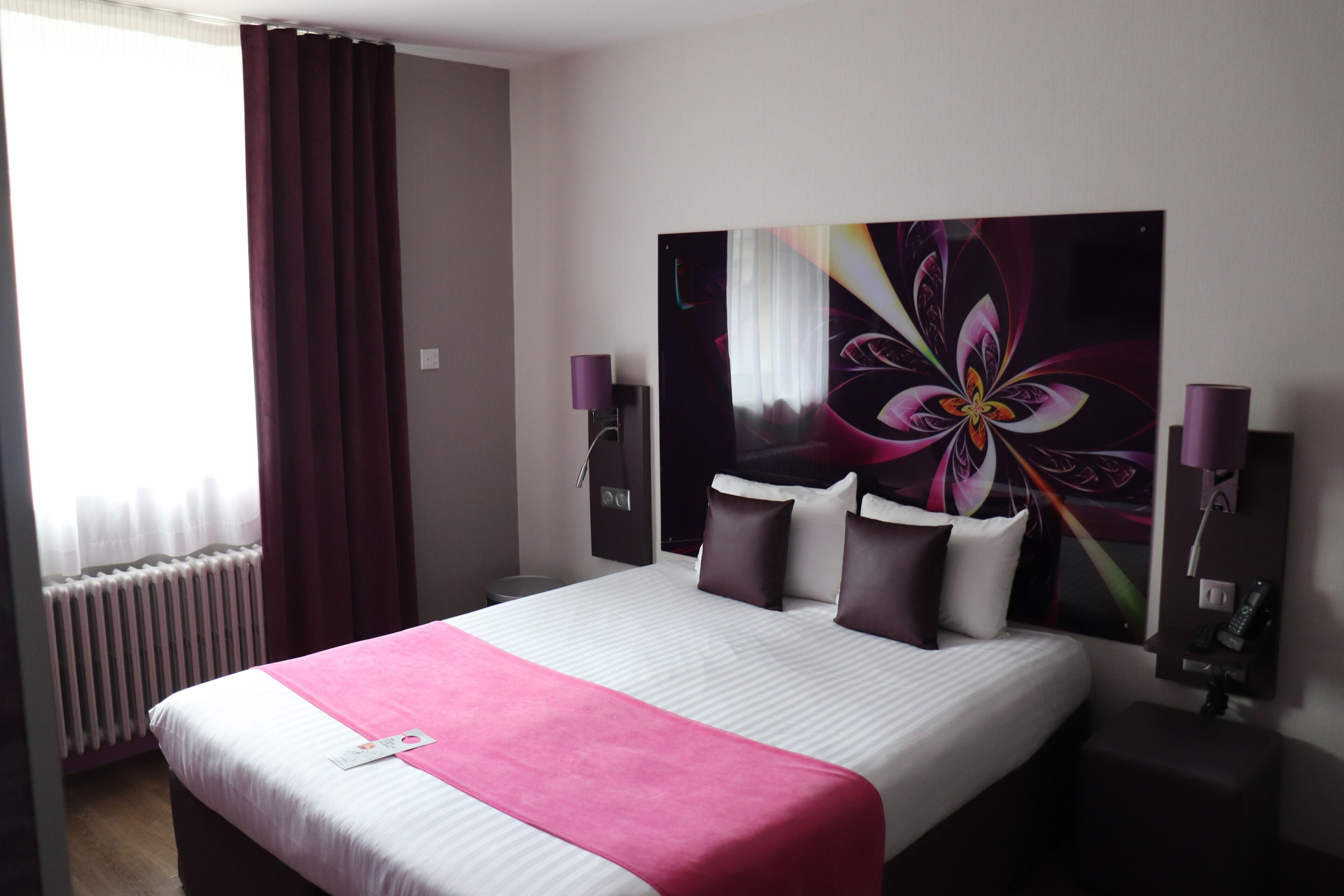 Une chambre d’hôtel avec un grand lit, une couvre-lit rose, une grande œuvre d’art florale accrochée au mur et une fenêtre avec des rideaux.
