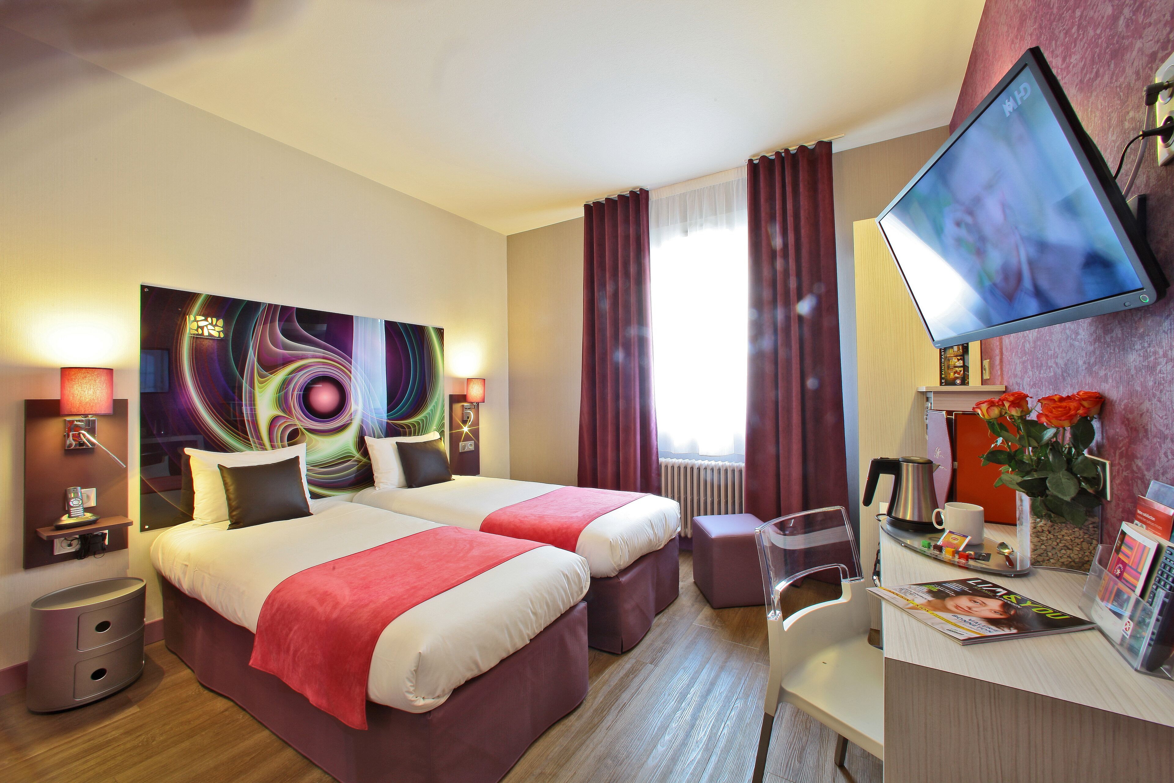 Hotelkamer met twee bedden, een bureau met een scherm, een koffiezetapparaat en een vaas met bloemen.