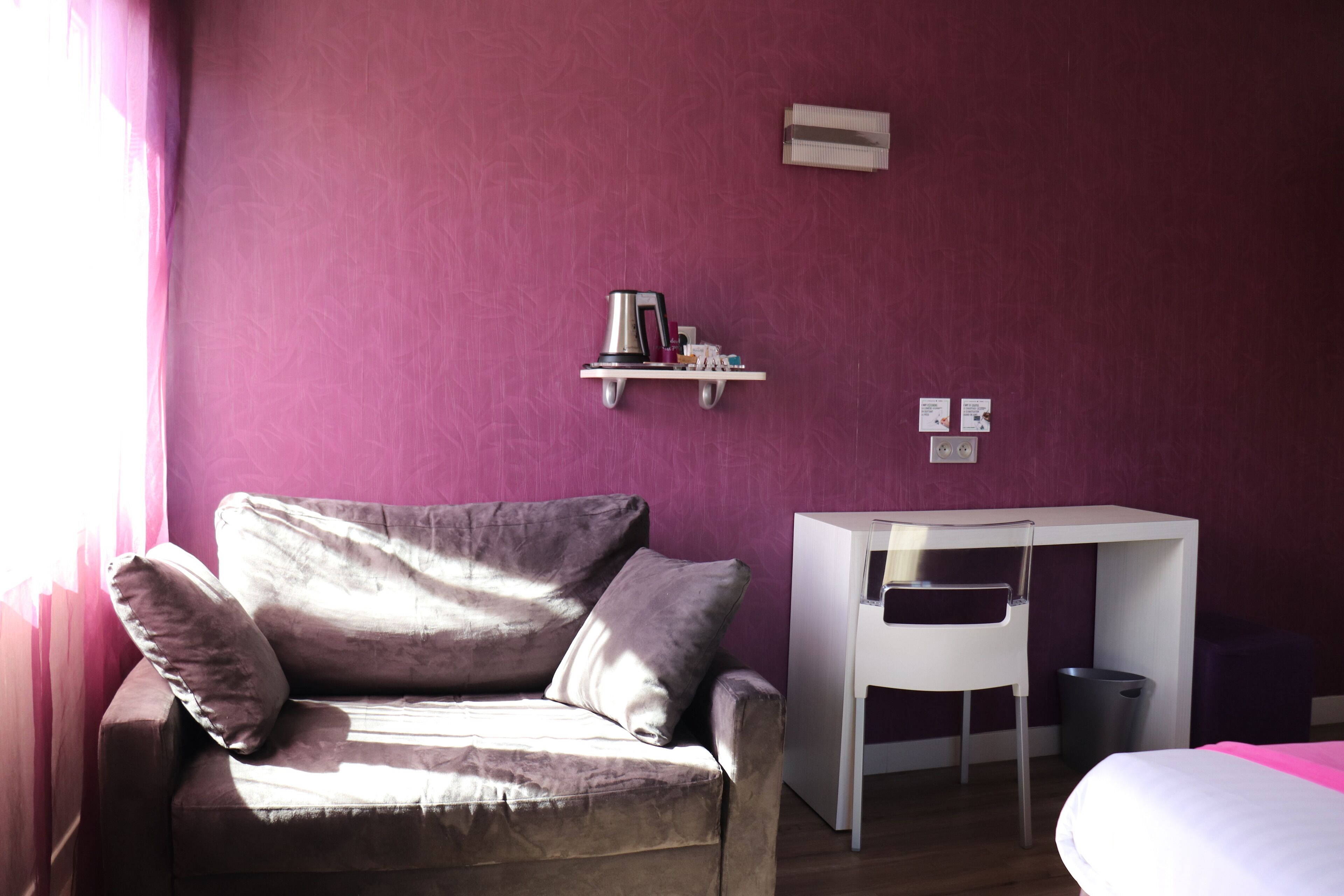 A room with a purple wall, a sofa with cushions, a small white desk, and a bed with a pink bedspread.
