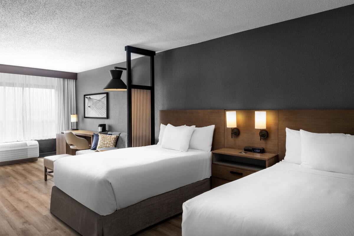Ein Hotelzimmer mit zwei Betten, einem Schreibtisch, einem Stuhl und einem großen Fenster mit Vorhängen.