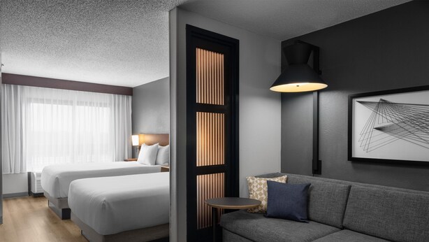 Ein Hotelzimmer mit zwei Betten, einem Schlafsofa, einem kleinen Tisch, einer Lampe und einem gerahmten abstrakten Kunstwerk an der Wand.