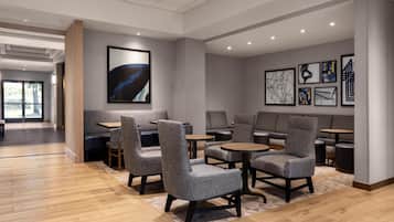 Een moderne lounge met grijze gestoffeerde fauteuils en tafels, kunstwerken in lijsten aan de muren, en een houten vloer.