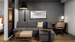 Ein modernes Hotelzimmer mit grauem Sofa, einem kleinen Holztisch, einem Schreibtisch mit Stuhl, einer schwarzen Stehlampe und einem gerahmten abstrakten Kunstwerk an der Wand.