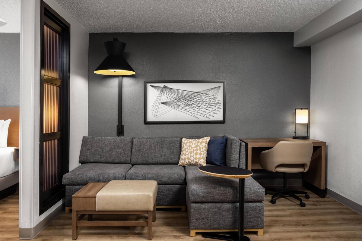 Ein modernes Hotelzimmer mit grauem Sofa, einem kleinen Holztisch, einem Schreibtisch mit Stuhl, einer schwarzen Stehlampe und einem gerahmten abstrakten Kunstwerk an der Wand.