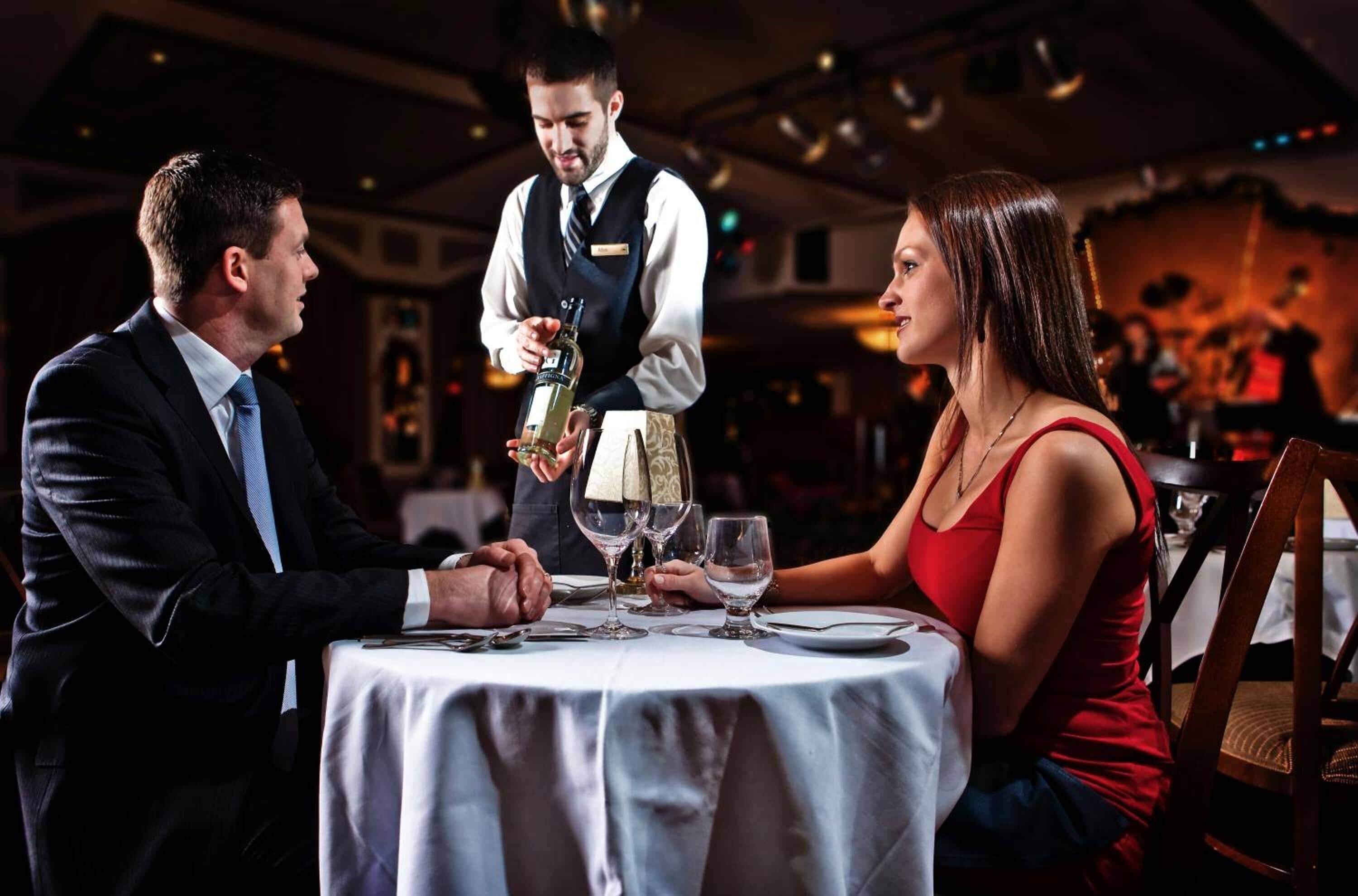 A formal dining setting with a waiter serving a bottle of wine to a couple at a round table.