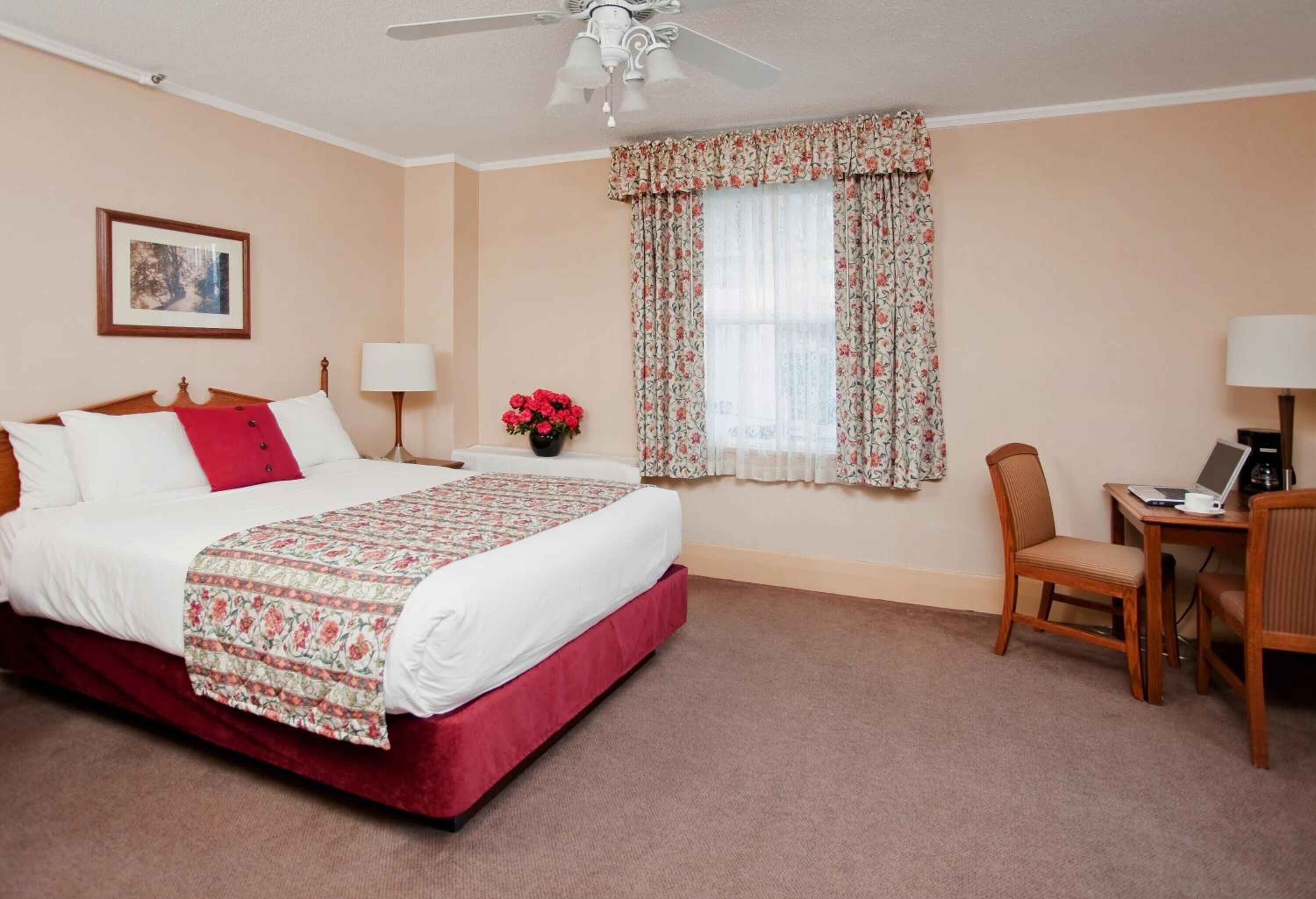 Habitación de hotel con cama, escritorio, silla, ventilador de techo, ventana con cortinas florales y un jarrón con flores.