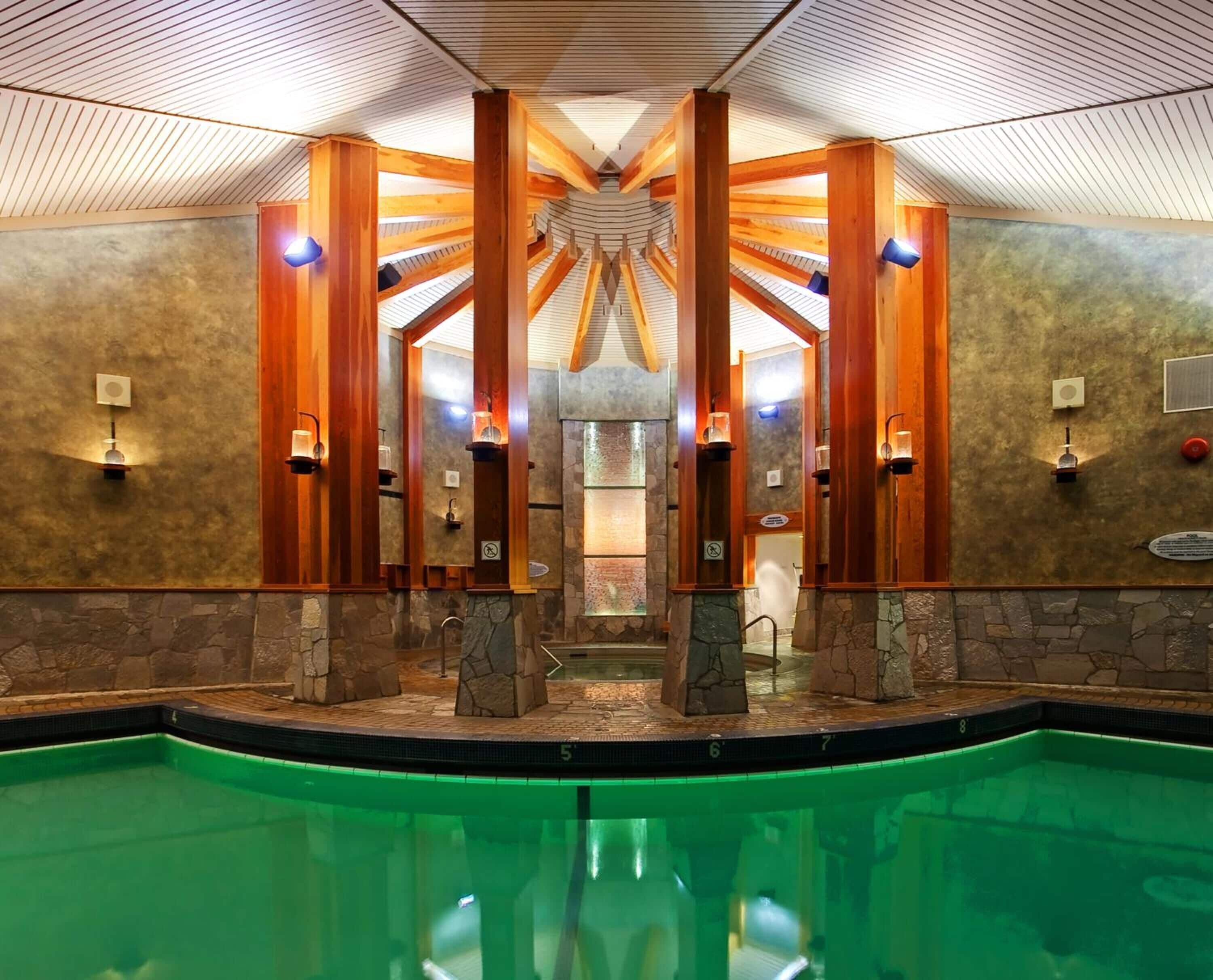 A modern indoor pool area with a central water feature, stone walls, and wooden beams.