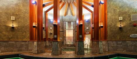 A modern indoor pool area with a central water feature, stone walls, and wooden beams.