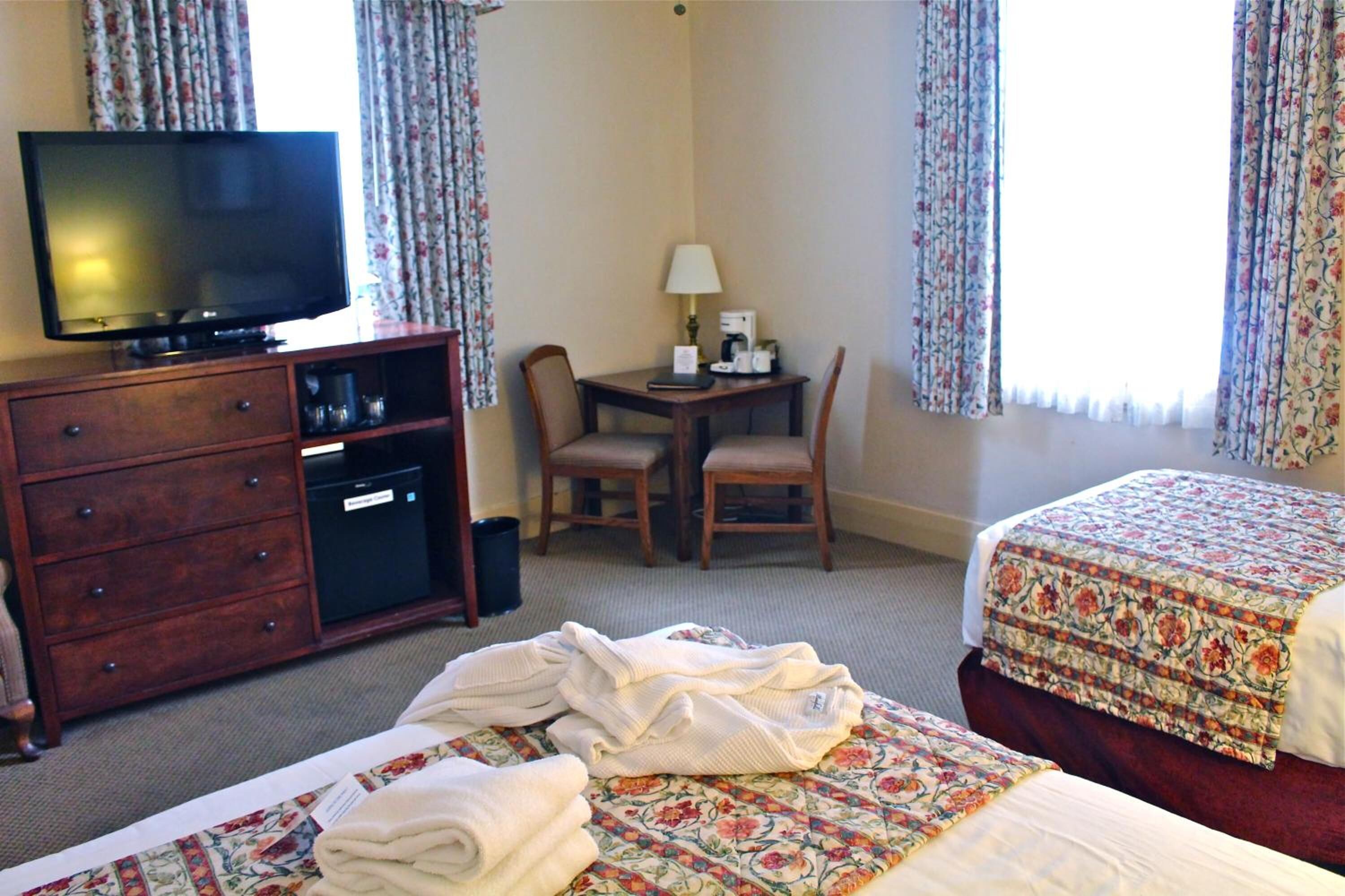 Habitación de hotel con televisión, escritorio con lámpara, dos sillas, cama con ropa de cama floral y cómoda.
