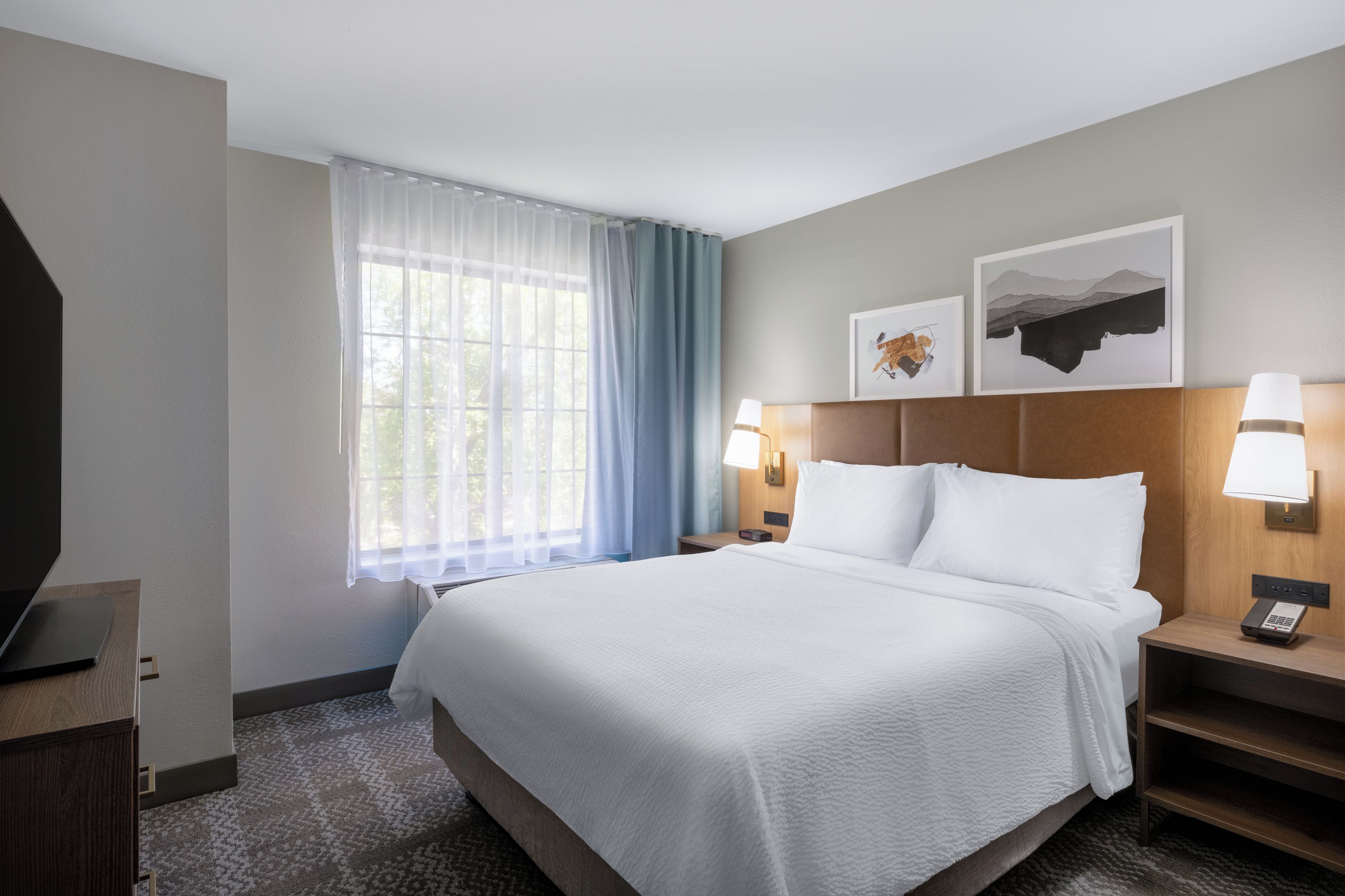 A hotel room with a large bed, two bedside tables with lamps, a flat-screen TV, and a window with sheer curtains.