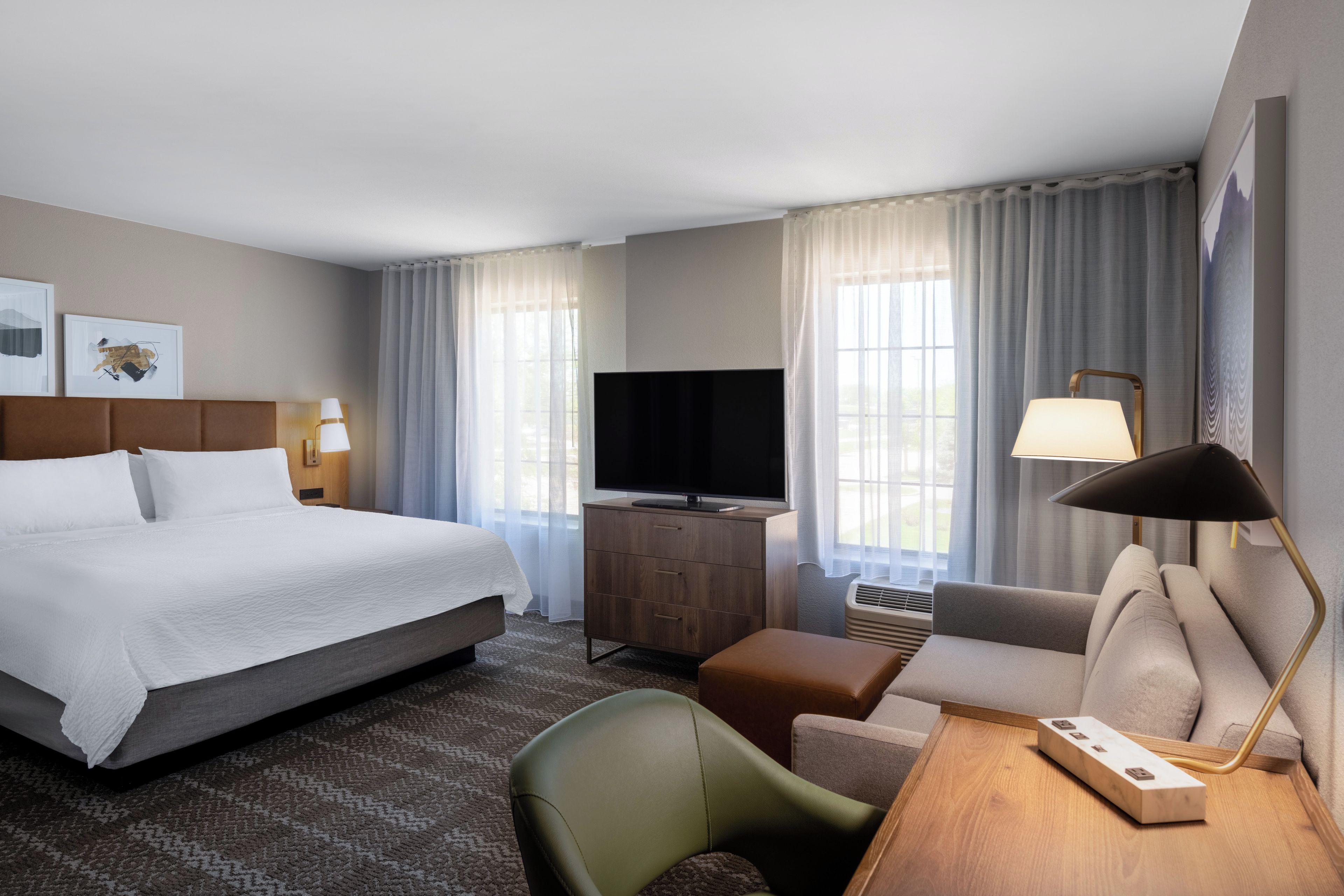 A hotel room with a bed, a desk with a lamp, a television, and a chair.