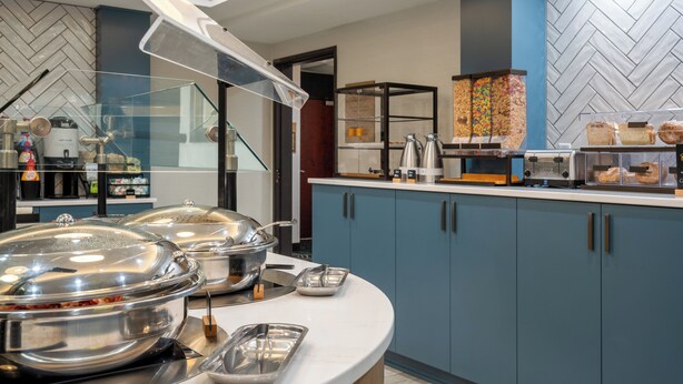 A buffet area with covered food stations, a coffee machine, and a display of pastries. - Staybridge Suites West Des Moines by IHG (West Des Moines)