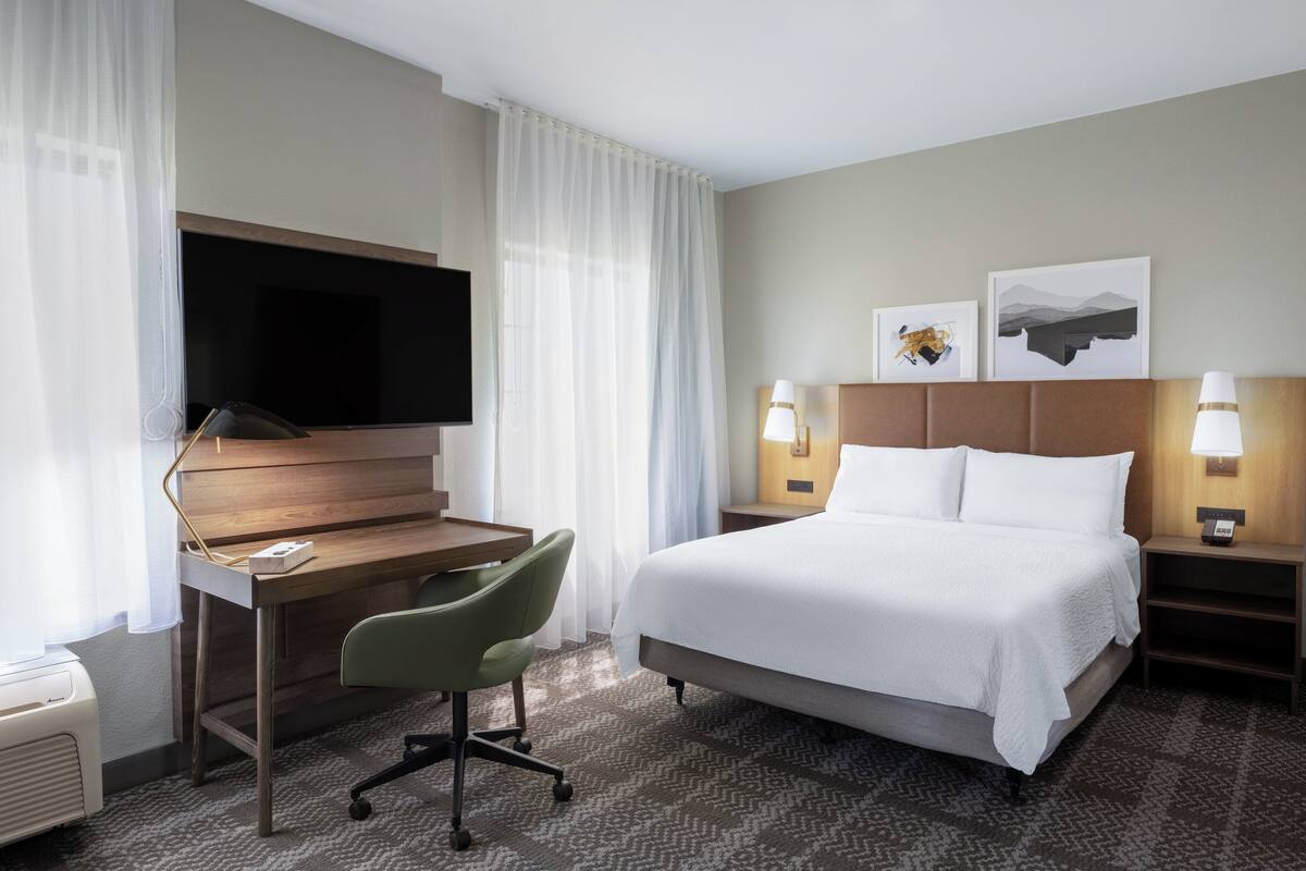 A hotel room with a bed, a desk with a chair, a television, two bedside lamps, and two framed pictures on the wall.
