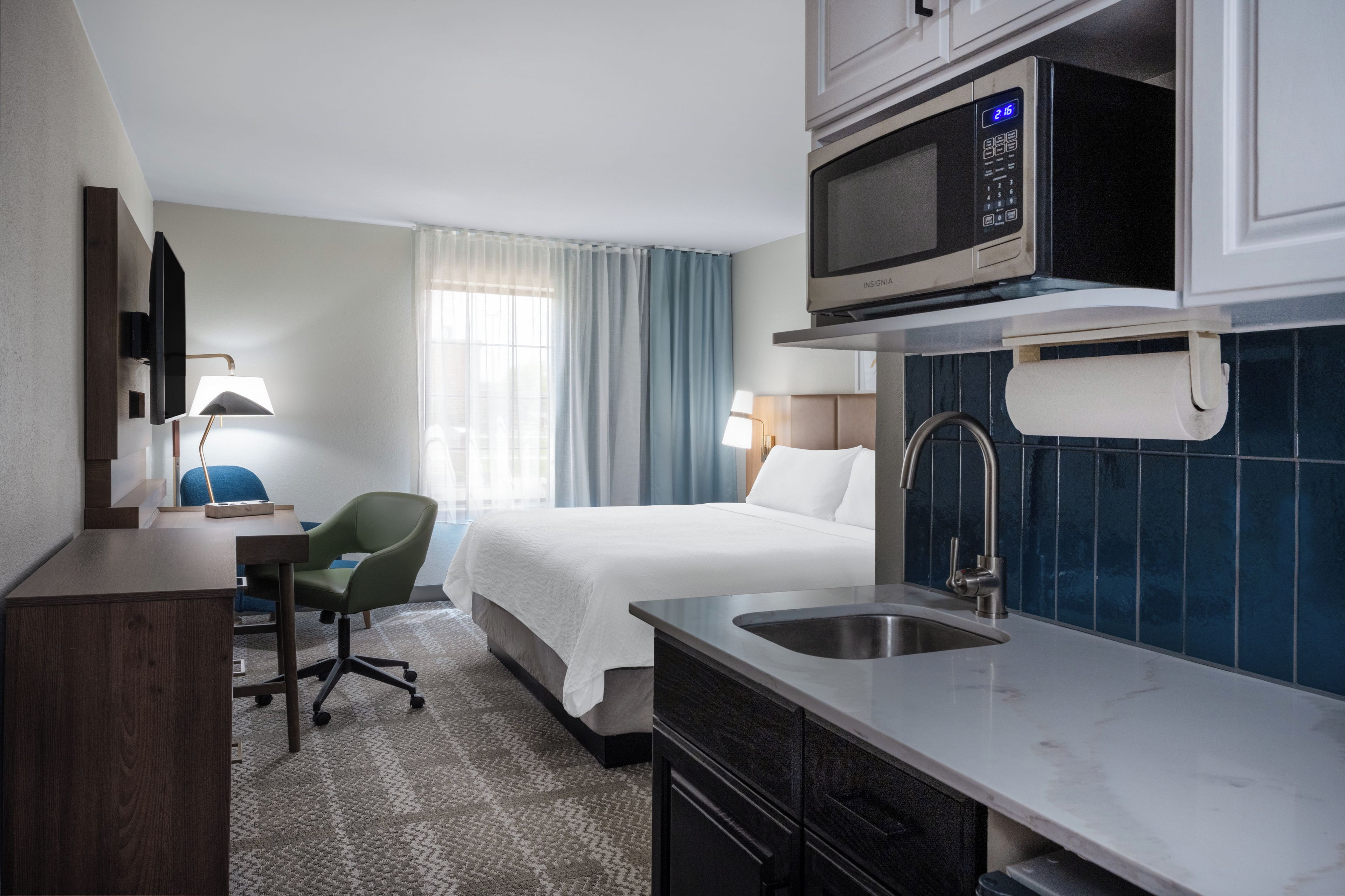 A hotel room with a bed, a desk, a chair, a microwave, and a sink.
