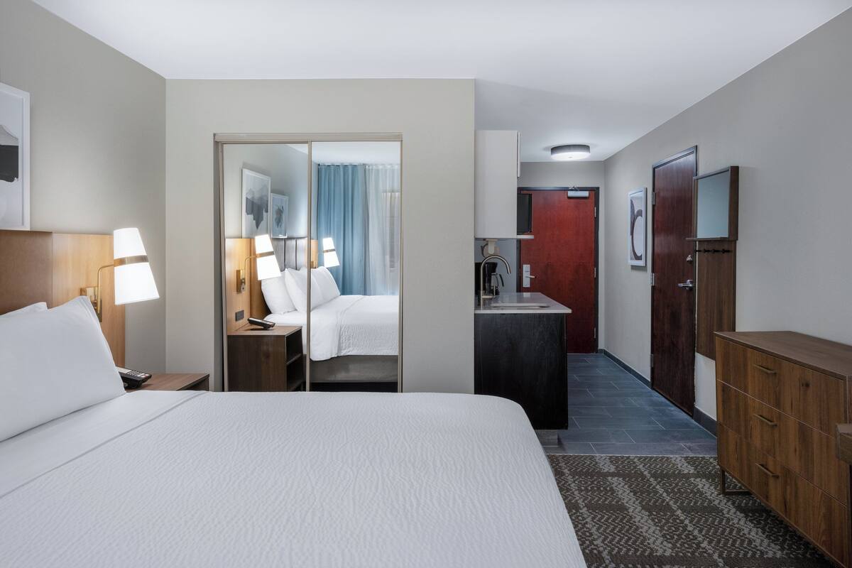 A hotel room with a large bed, a desk with a lamp, a wardrobe, a mirror, and a door leading to a bathroom.