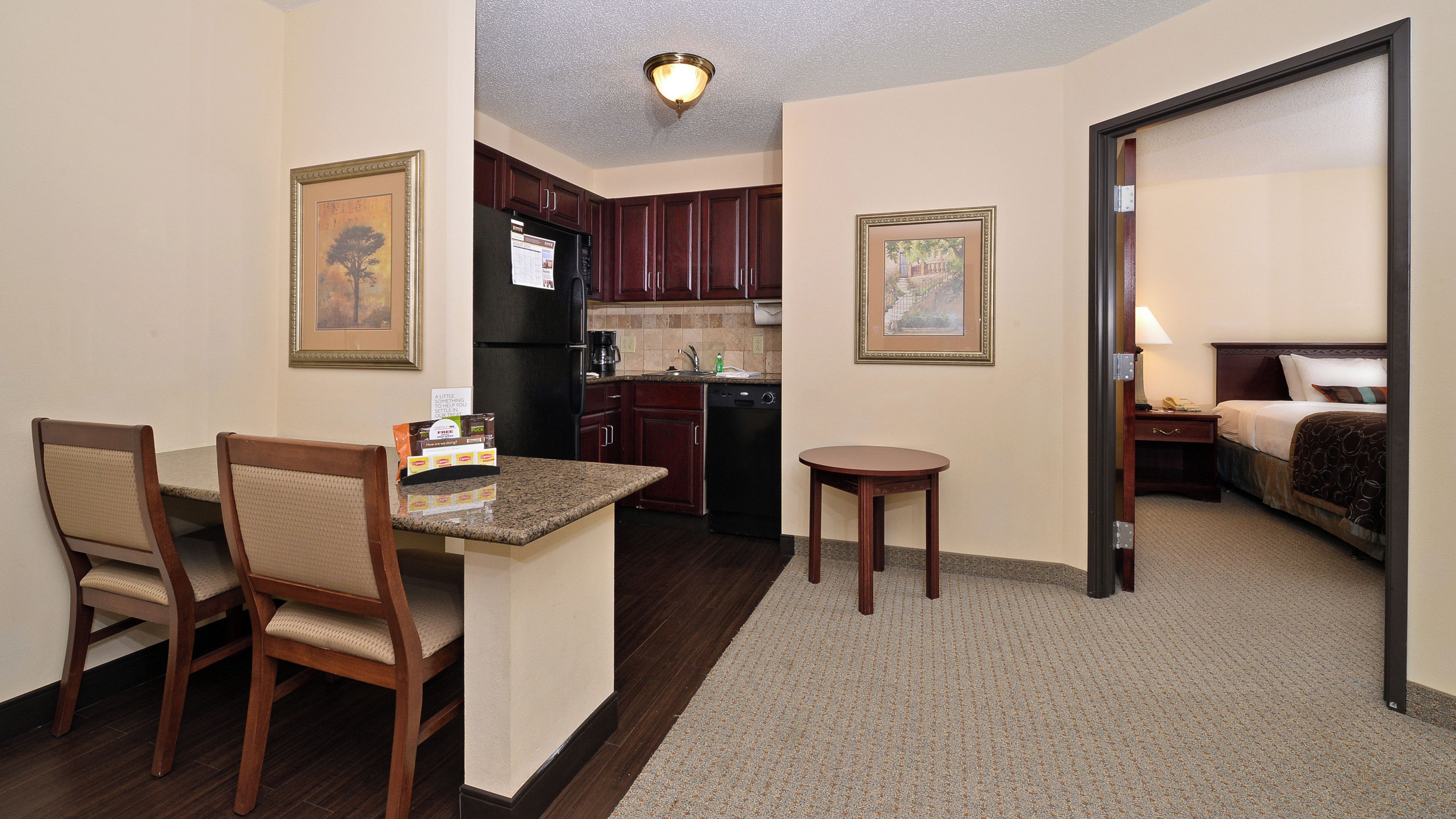 Habitación de hotel compacta con kitchenette, un comedor con dos sillas, una mesita y una cama con mesita de noche.