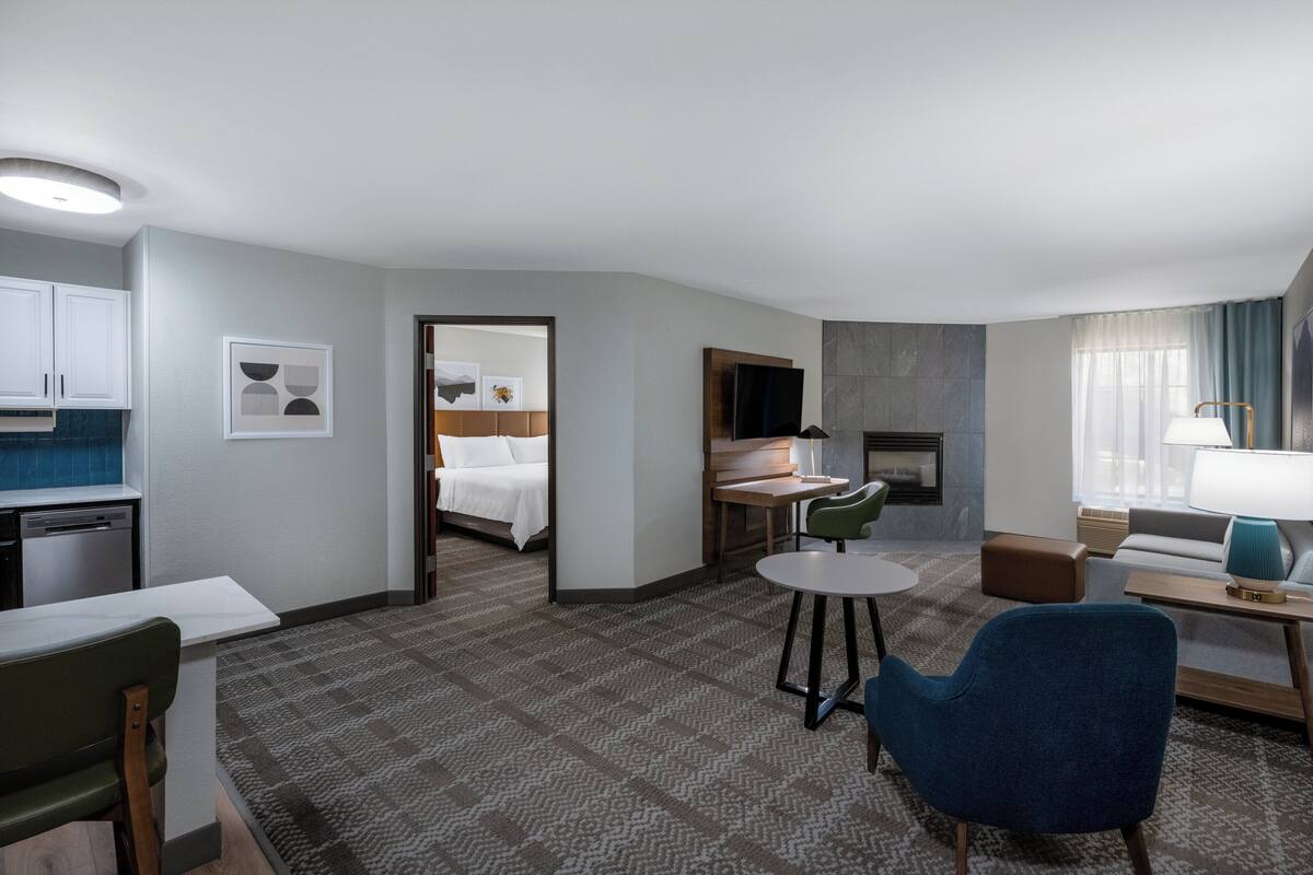 A hotel room with a kitchenette, a dining area, a living area with a sofa and armchair, a bed, and a fireplace.