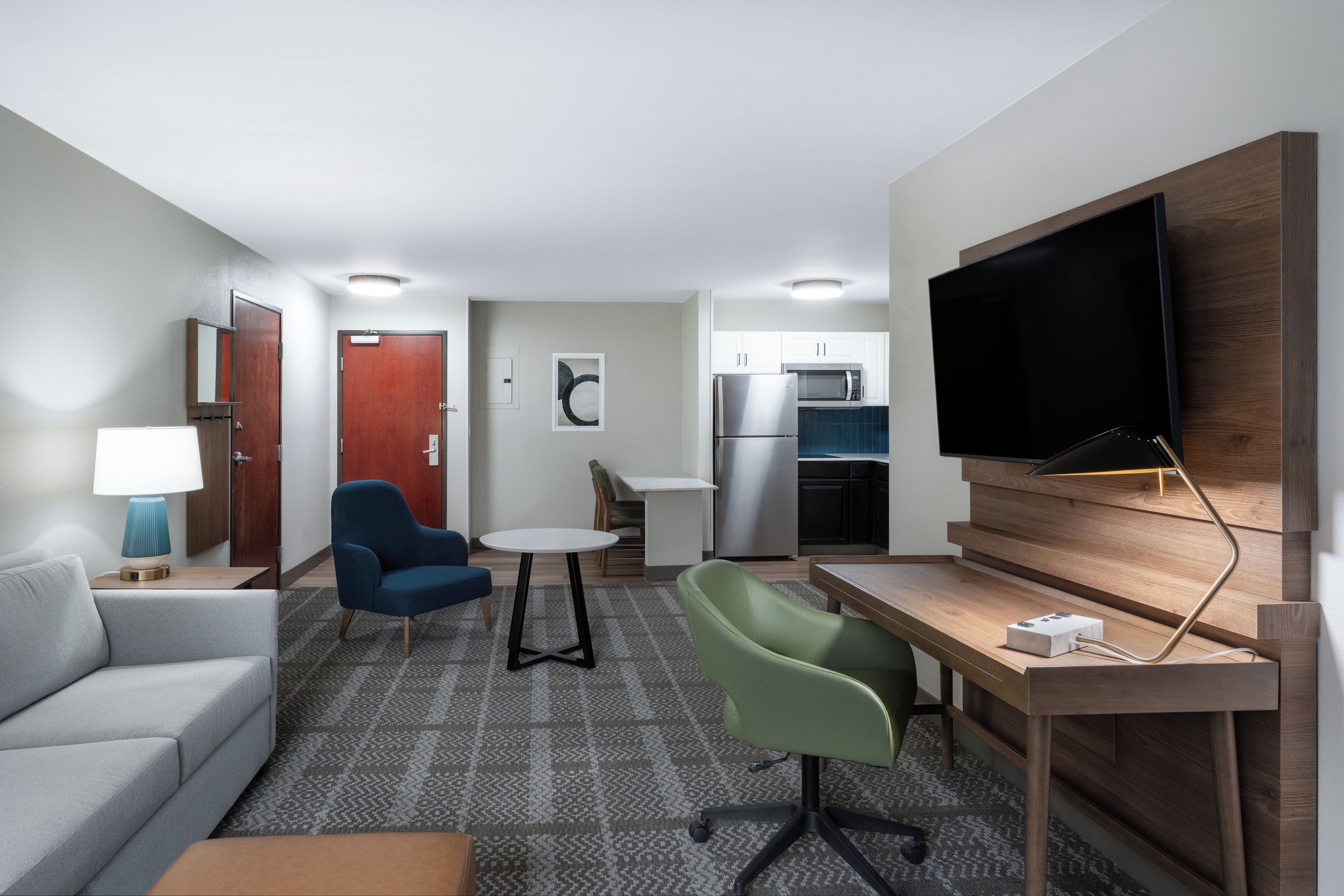 A compact hotel room with a kitchenette, a flat-screen TV mounted on the wall, a sofa, a dining table with chairs, and a patterned carpet.