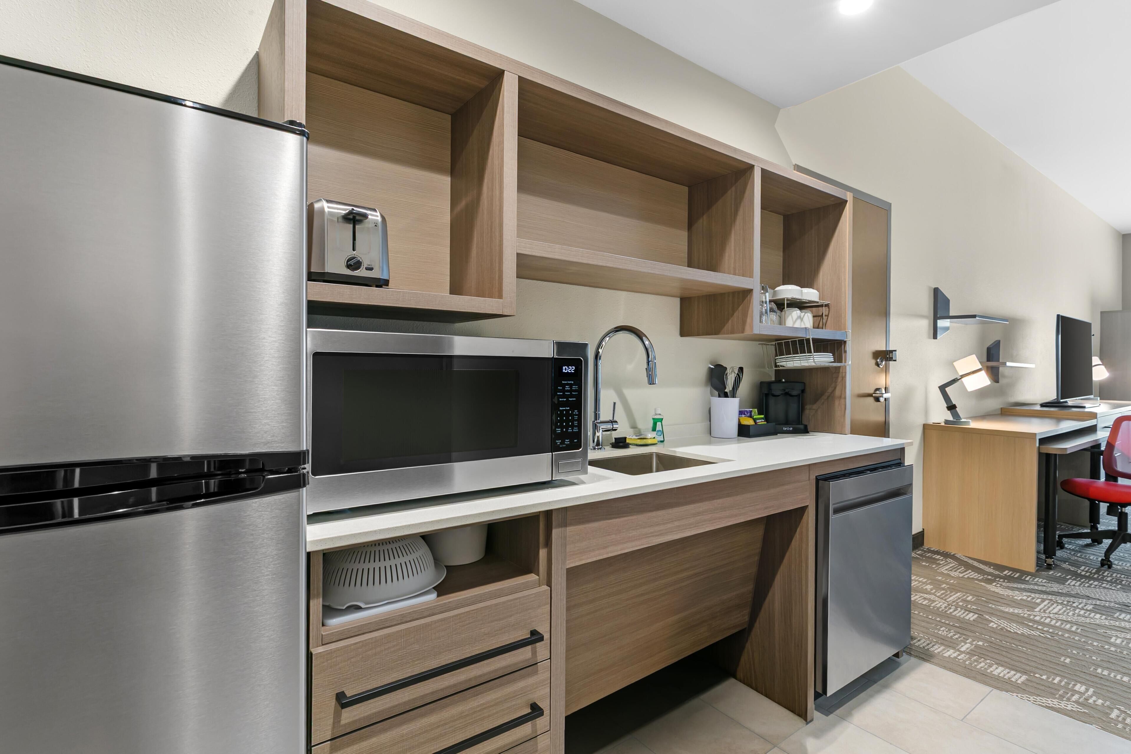 A modern kitchen with a stainless steel refrigerator, microwave, and dishwasher, wooden cabinetry, and a small workspace with a desk and chair.
