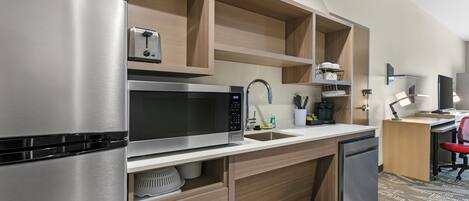 A modern kitchen with a stainless steel refrigerator, microwave, and dishwasher, wooden cabinetry, and a small workspace with a desk and chair.