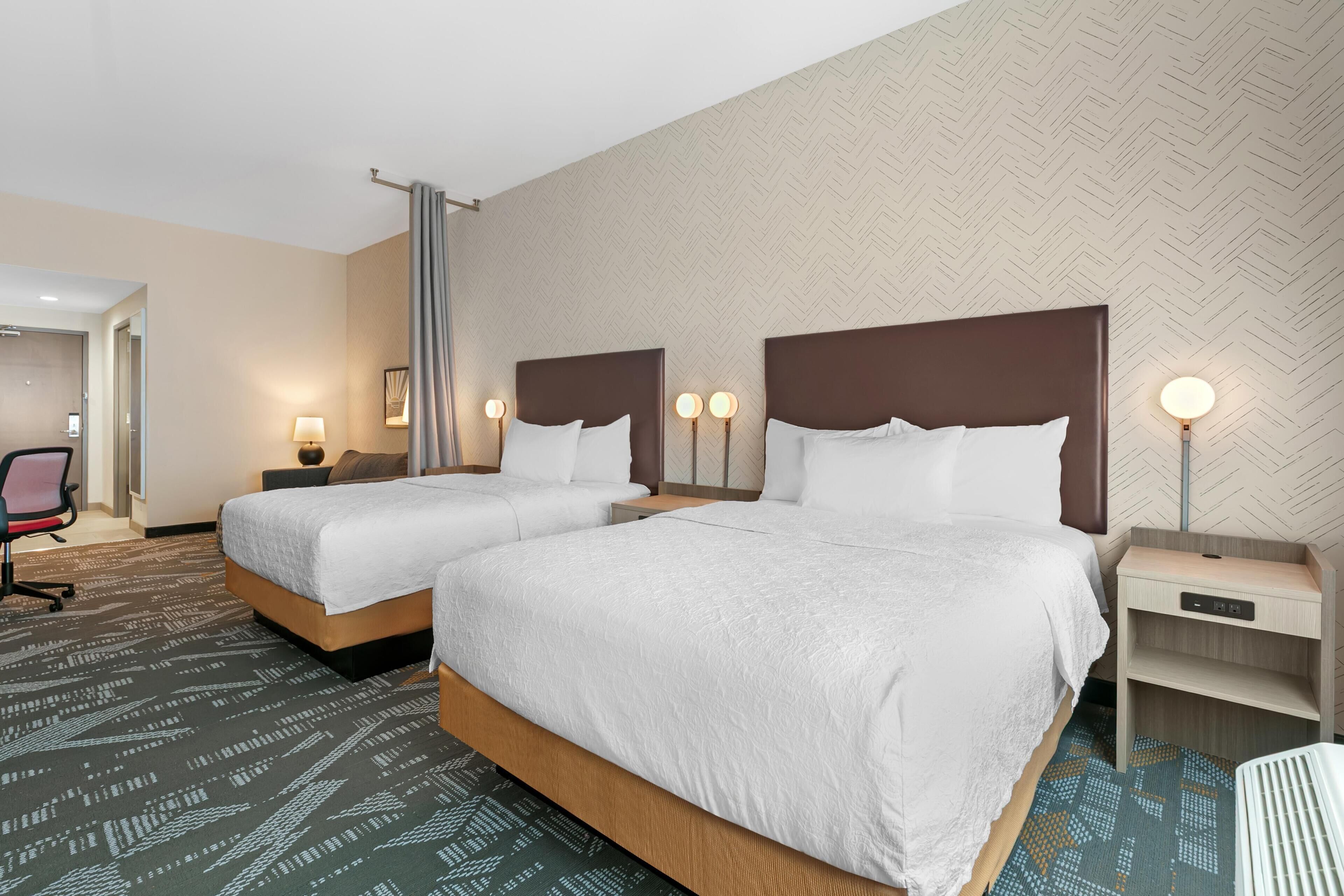 A hotel room with two beds, a desk, a chair, and a nightstand with lamps.