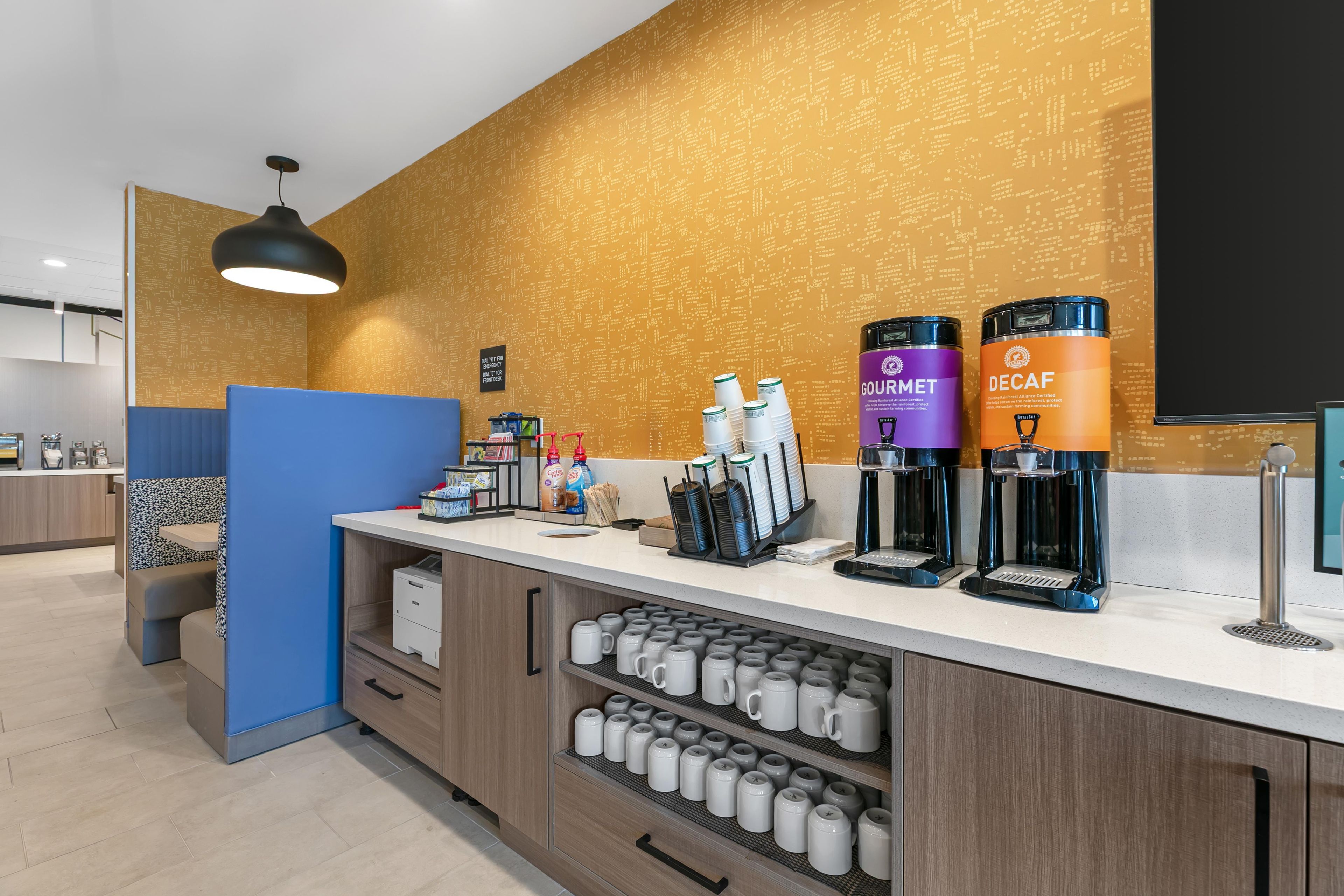 A modern coffee station with branded coffee dispensers, a counter with various coffee-making equipment, and a seating area with a blue accent wall.