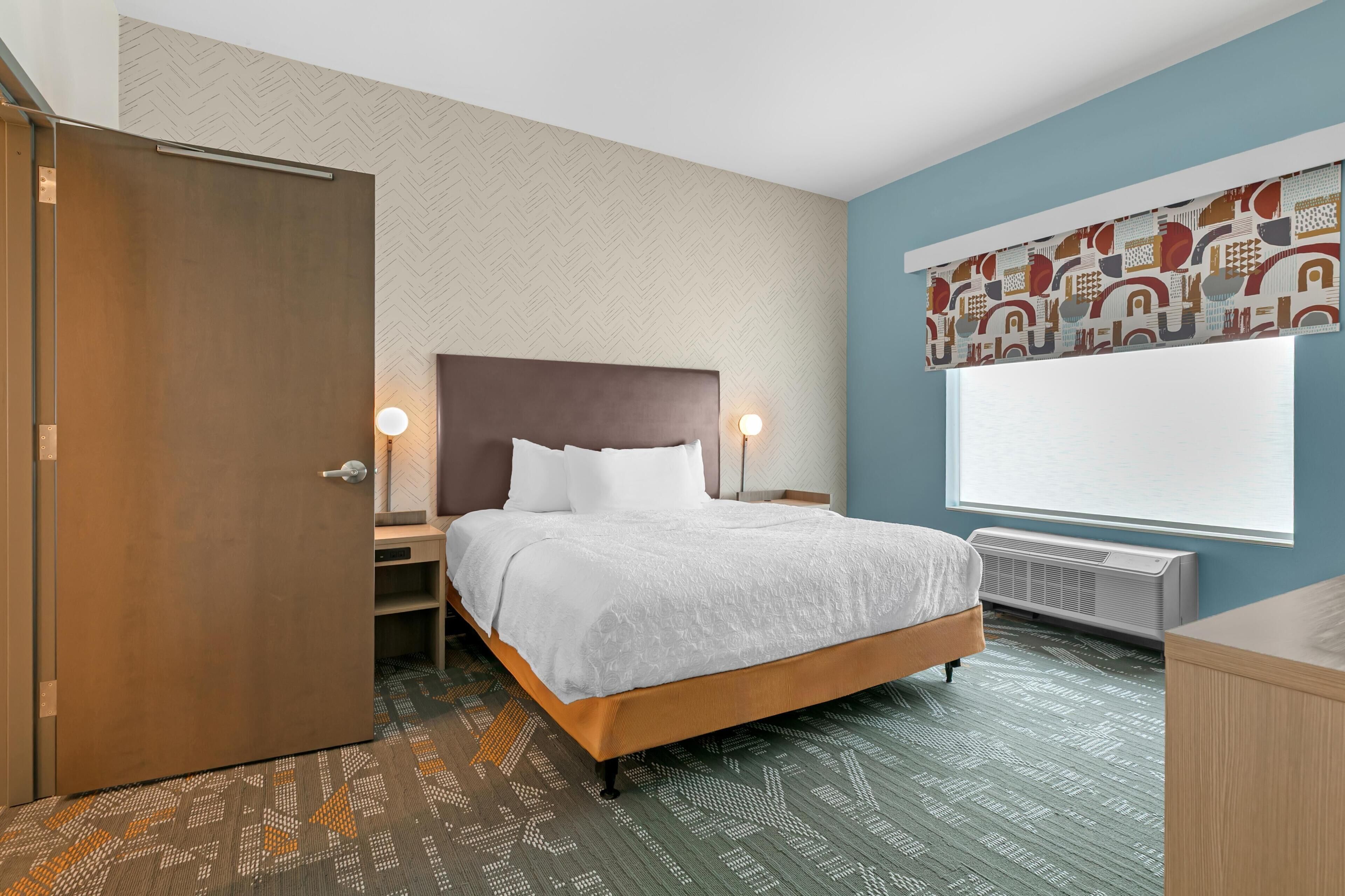 Habitación de hotel con cama, mesita de noche, lámpara, ventana con persianas, radiador y alfombra estampada.