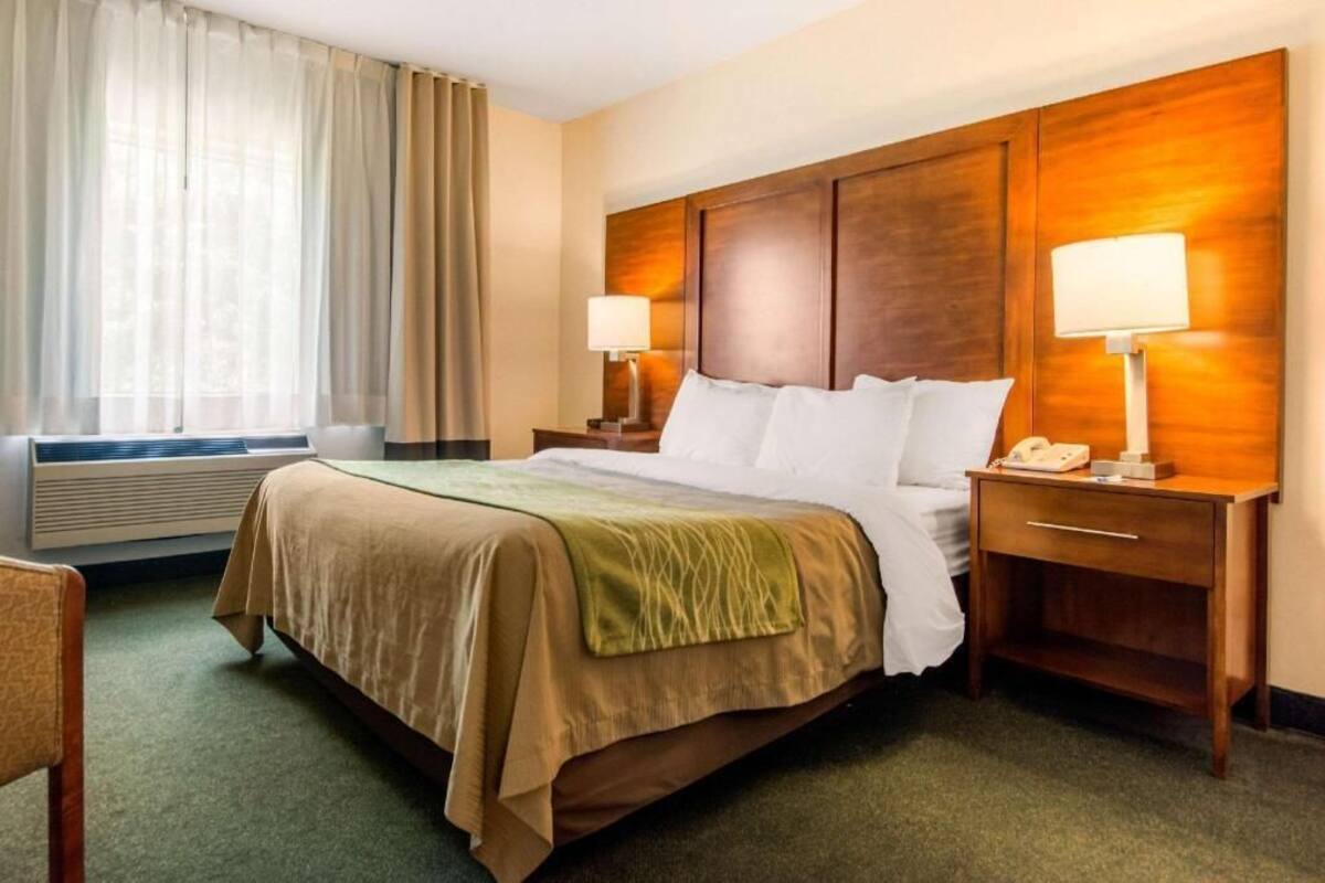 A hotel room with a bed, two bedside tables with lamps, a chair, a window with curtains, and a wall-mounted heater.