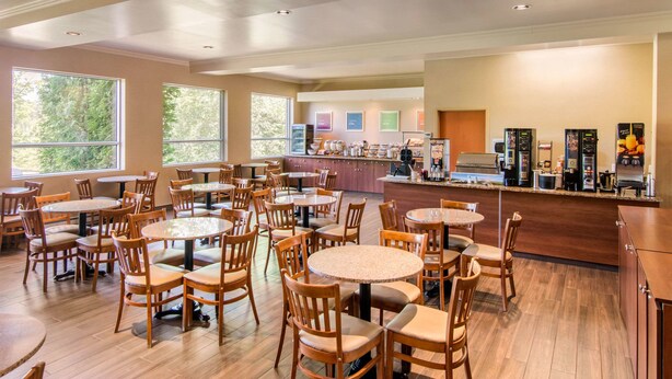 A spacious dining area with multiple round tables and wooden chairs, a kitchen area with a coffee station, and large windows providing natural light. - Comfort Inn & Suites Shawinigan (Shawinigan)