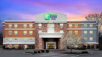 Welcome to the Holiday Inn Express & Suites, featuring a central entrance with double doors and a parking area surrounded by trees and shrubs.