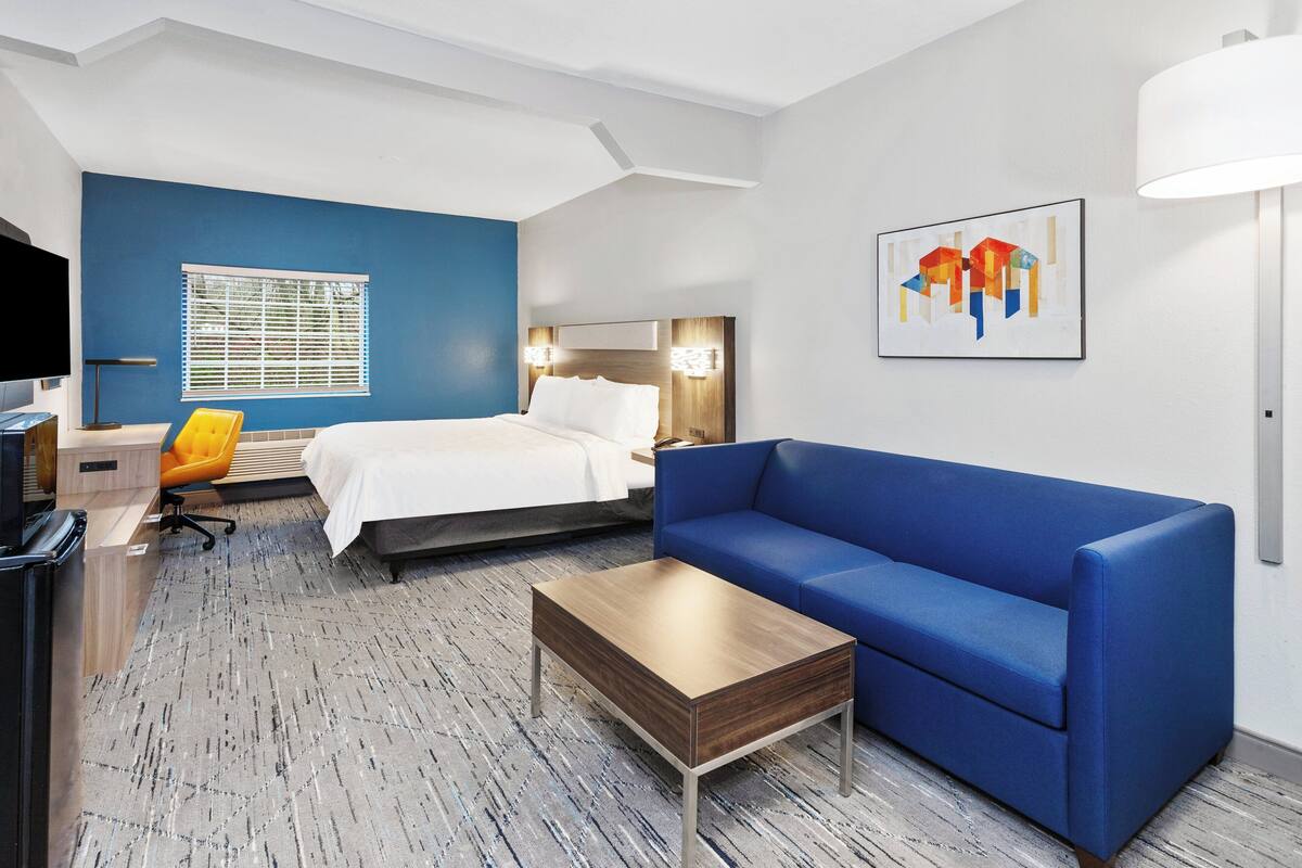 A hotel room with a bed, a blue sofa, a wooden coffee table, a desk with a chair, a TV, and a window with blinds.