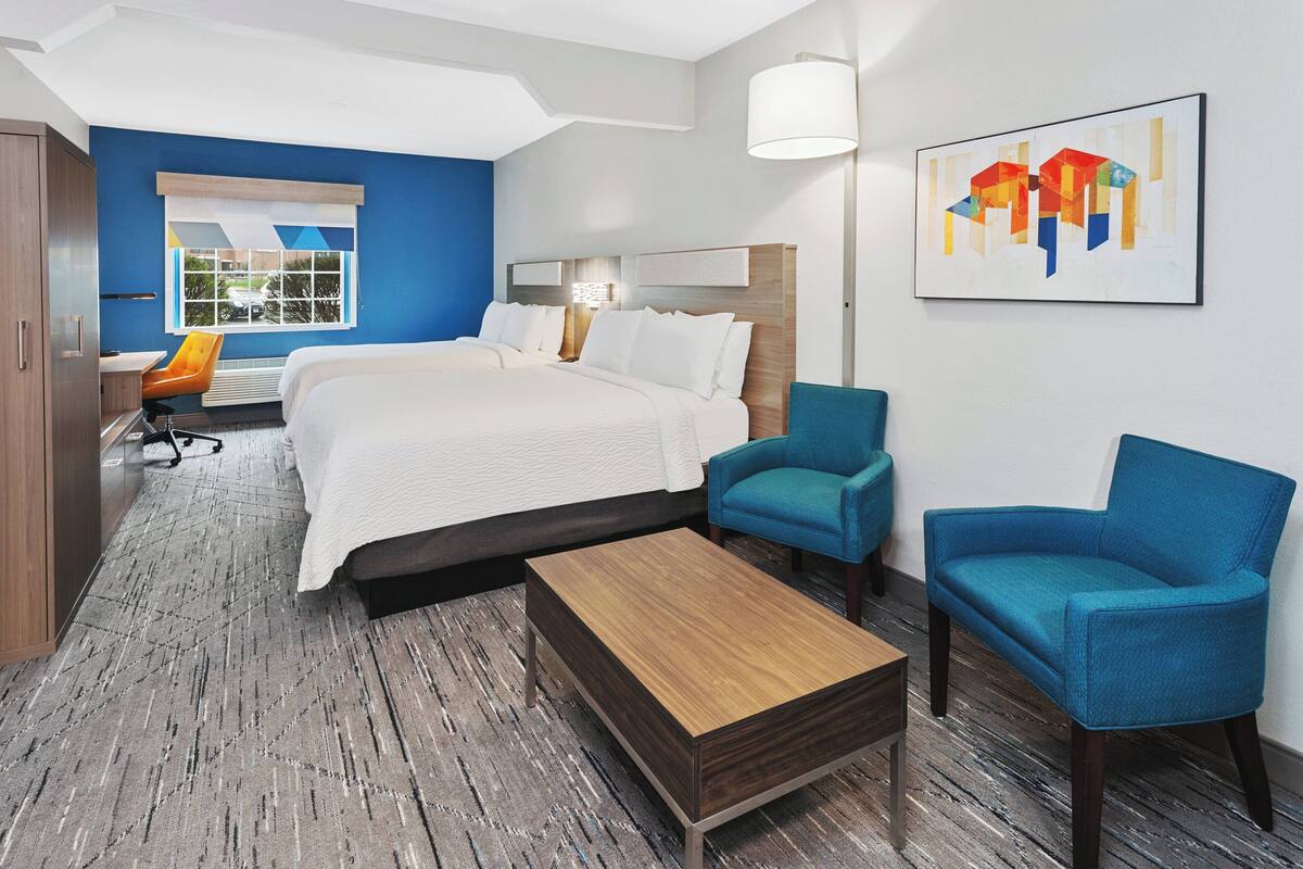 A hotel room with a large bed, two blue armchairs, a wooden coffee table, and a desk with an orange chair.