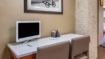 A hotel room with a desk, a computer, a keyboard, a mouse, and a framed picture on the wall.