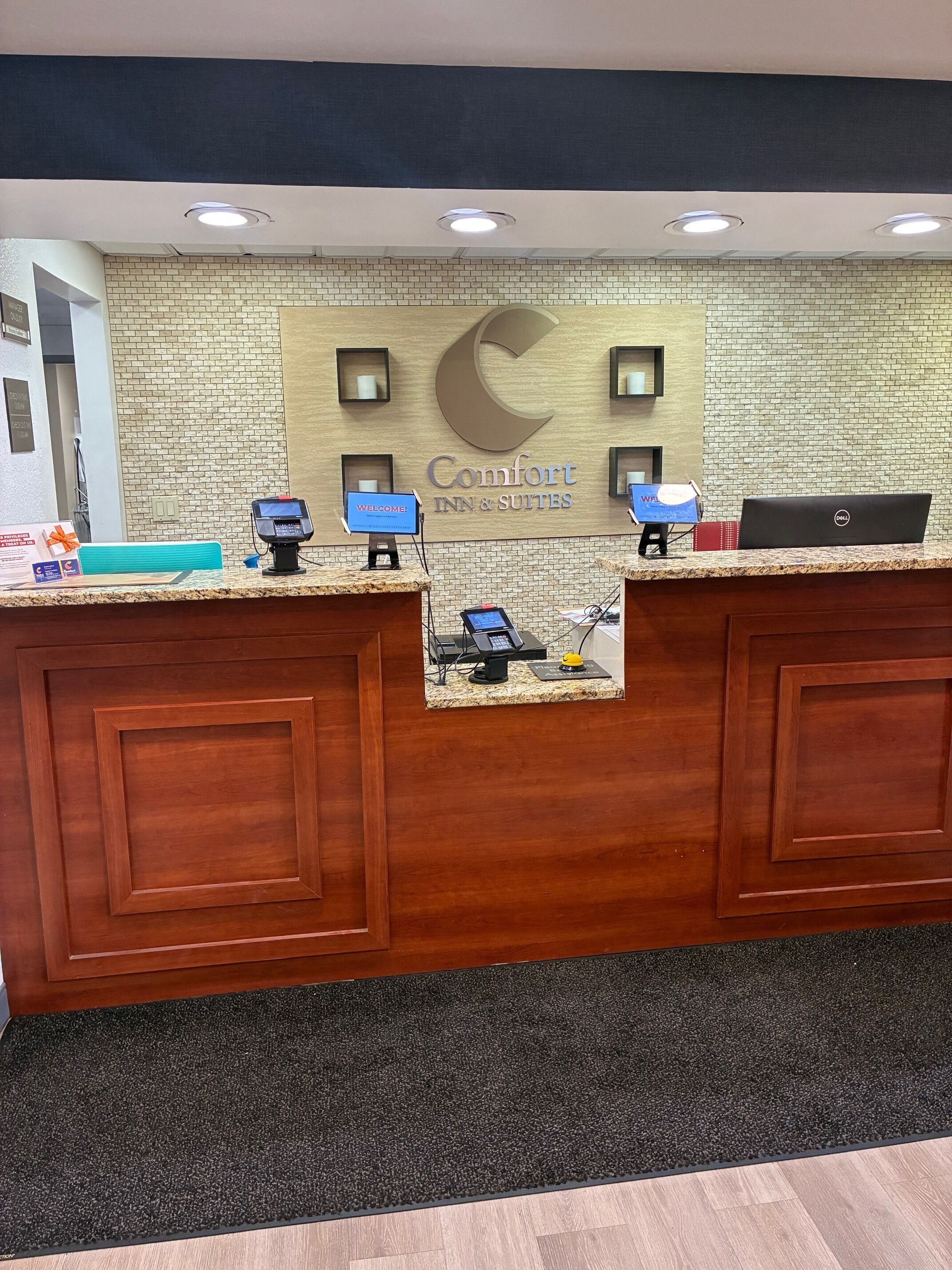 Una recepción de hotel con un muro de ladrillo, un letrero grande de 'Comfort Inn & Suites' y una laptop Dell en el mostrador.